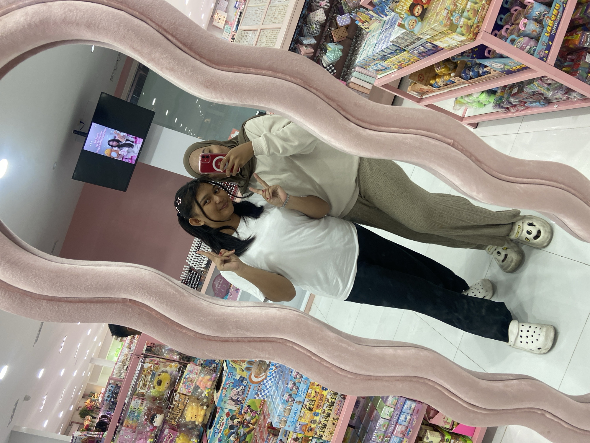Two young women are taking a mirror selfie in what appears to be a store. The woman on the left has dark hair pulled back and is wearing a white t-shirt and black pants, holding up two fingers in a peace sign. The woman on the right is wearing a hijab and a light-colored sweater over beige pants, holding her phone to take the photo and also making a peace sign with her fingers. The store has shelves stocked with various toys and merchandise, and a television screen is visible in the background. The mirror they are using is large and has a pink, plush, wavy frame. The floor is tiled. The overall setting suggests a retail environment, possibly a toy store or a store selling novelty items.