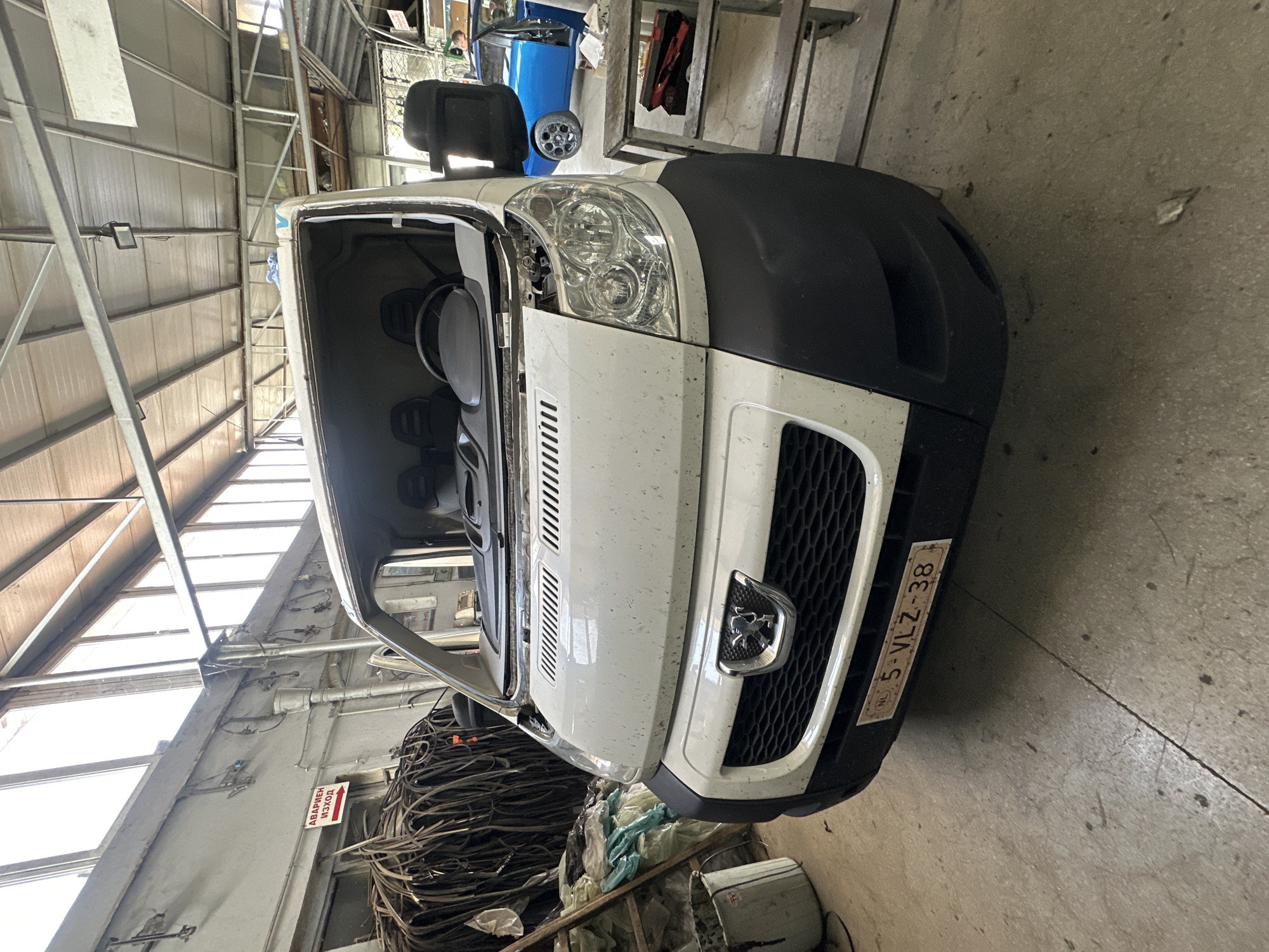 The image shows the front of a white van with its windshield removed, parked indoors in what appears to be a workshop or garage. The van has a black bumper and a honeycomb grille with a silver Peugeot logo. A license plate with the text "NL 5-VLZ-38" is visible on the bumper. To the left of the van, there is a pile of dark-colored hoses and some plastic sheeting. A white bucket containing a dark substance is also visible. In the background, a blue sports car is parked, and a person can be seen in the distance. The environment has a concrete floor and a metal roof with exposed beams and lighting fixtures. A sign with Cyrillic text and a red arrow pointing to the right is mounted on the wall in the upper left corner. The overall scene suggests vehicle repair or maintenance.