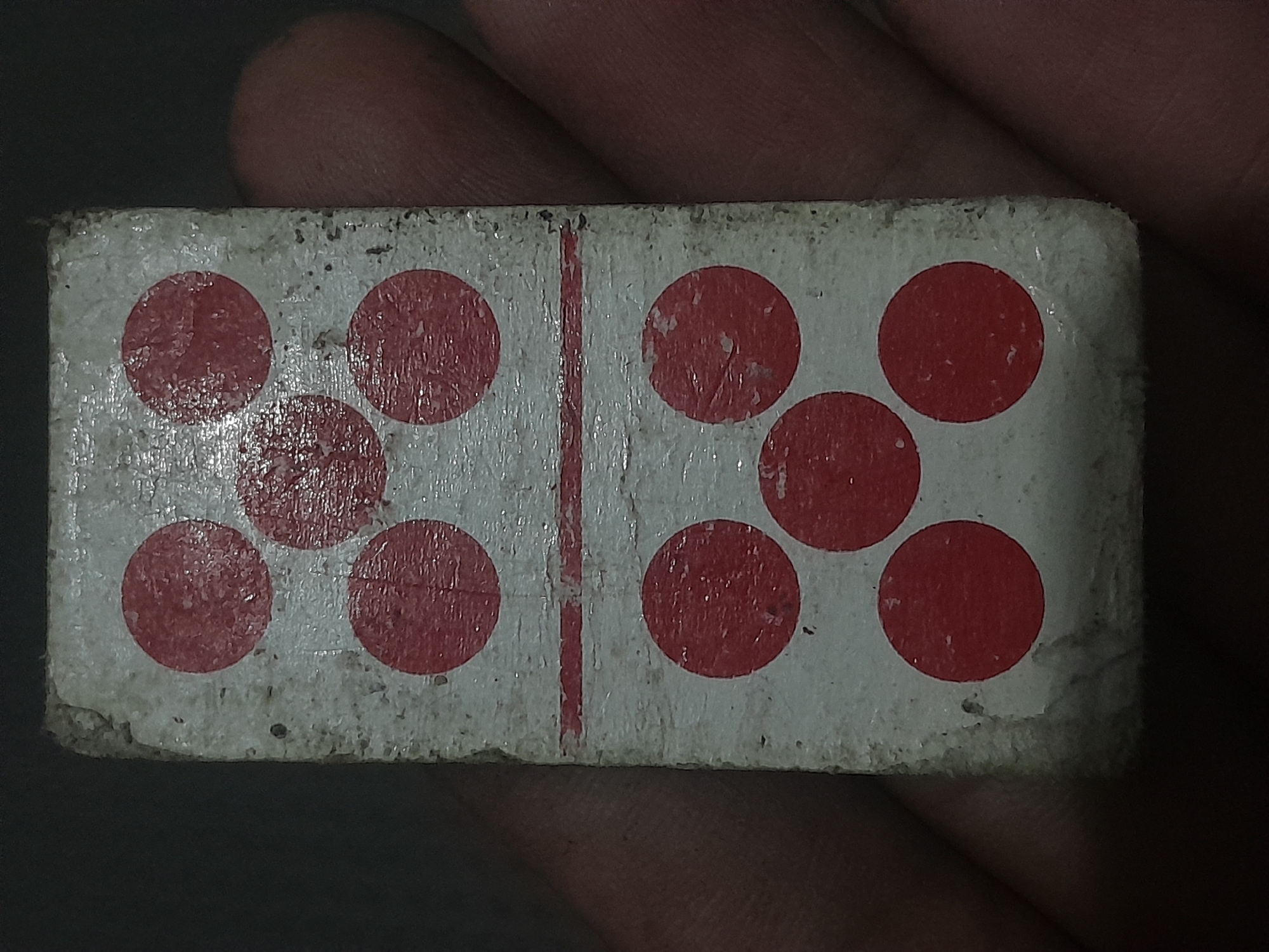 The image shows a close-up, top-down view of a domino with a white background divided by a horizontal red line. The top half of the domino displays four red dots arranged in a diamond pattern. The bottom half of the domino also displays four red dots, arranged in a similar diamond pattern, but with an additional red dot in the center. The domino appears worn and aged, with scuffs and chipped edges visible on its surface. It is being held by a person's hand, with fingers and palm visible on the right side of the frame. The background is dark and out of focus, suggesting the photo was taken in a dimly lit environment. There is no discernible information or context within the image that would indicate its location in Depok, Indonesia.