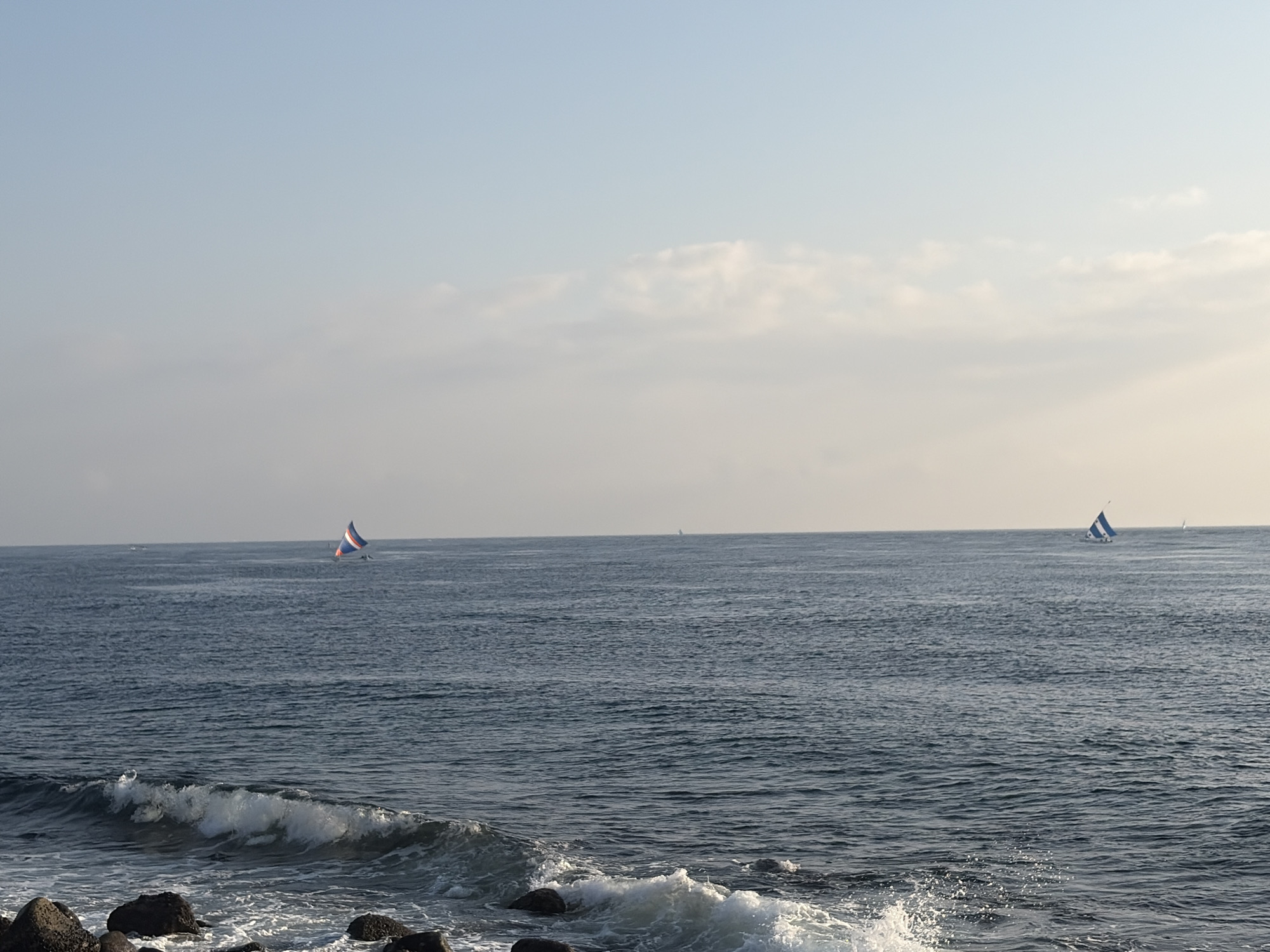 This is a wide shot of the ocean at sunset. The sky is a pale blue with some wispy clouds. The ocean is a deep blue, with small waves breaking on the shore. Two sailboats are visible in the distance. The foreground shows a rocky shoreline with waves crashing against the rocks. The scene is serene and peaceful. The location is the coast of Abang, Indonesia.