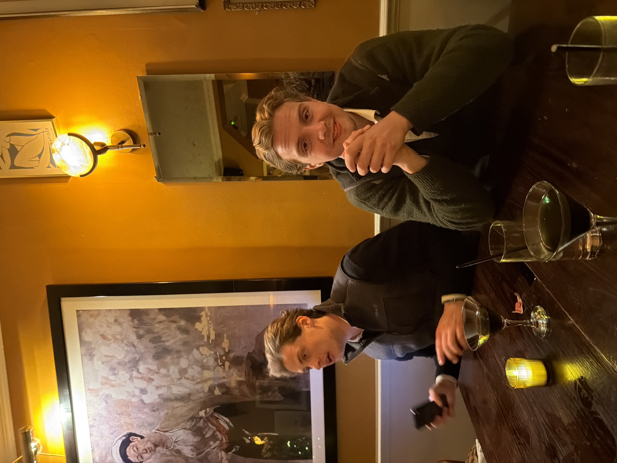 Two young men are sitting at a dark wooden table in a dimly lit restaurant. The man on the right, closest to the viewer, wears a dark green sweater and smiles at the camera while clasping his hands. To his right, on the table, there are empty glasses. The man on the left, in the background, wears a dark vest over a light shirt and is looking down at his phone. On the table in front of him, there are drinks and a lit candle.
Behind the man in the sweater is a framed mirror reflecting part of the restaurant. Behind the other man is a large framed painting. On the wall, there is a wall sconce with a spherical light fixture and a framed print with a Matisse drawing. Given the style of the restaurant and the artwork visible, it is possible that the location is in London, United Kingdom.