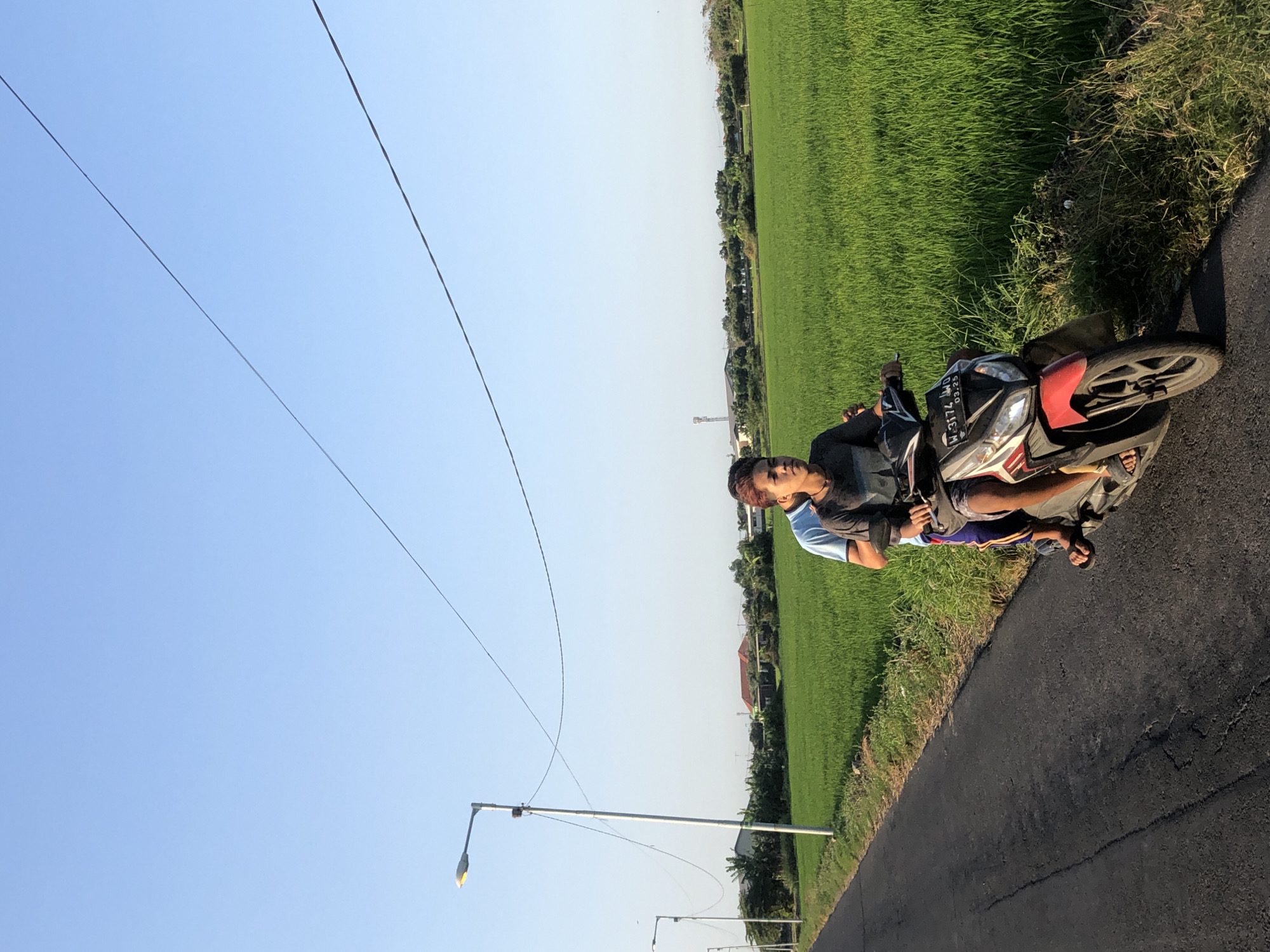 Two individuals are riding a red and white motorcycle on an asphalt road. The person in front is driving, wearing a dark gray t-shirt and blue shorts, with dark hair. The person behind them is wearing a light blue t-shirt. They are riding next to a lush green rice field. In the background, there are trees, houses, and a utility pole with streetlights. Power lines are visible stretching across the clear blue sky. The license plate on the motorcycle reads "W-3174-JW". The scene appears to be in Kalitengah, Indonesia, during daylight hours.