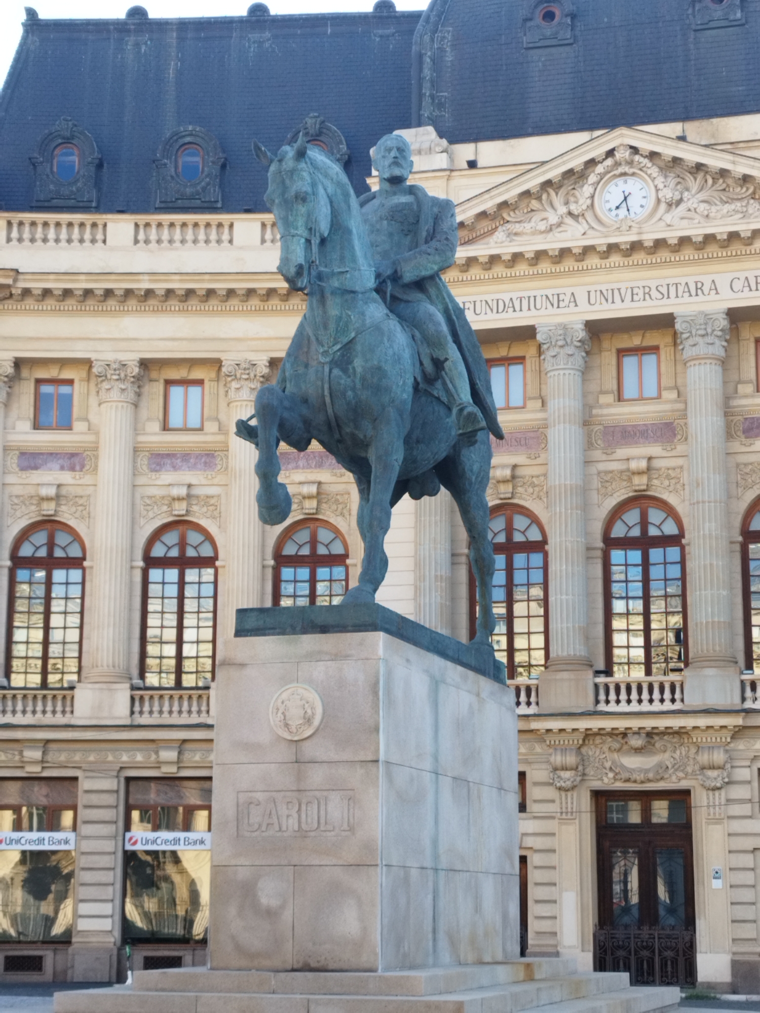 The image shows a bronze equestrian statue of Carol I, King of Romania, positioned in front of the Palace of the University Foundation, located on Bulevardul Regina Elisabeta in Bucharest, Romania. The statue depicts Carol I on horseback, facing left. The monument stands on a stone pedestal inscribed with "CAROLI I". Behind the statue, the neoclassical architecture of the palace is visible, featuring ornate columns, arched windows, and a large clock on the facade. The building also displays the inscription "FUNDATIUNEA UNIVERSITARA CAROL I". The sky is overcast.