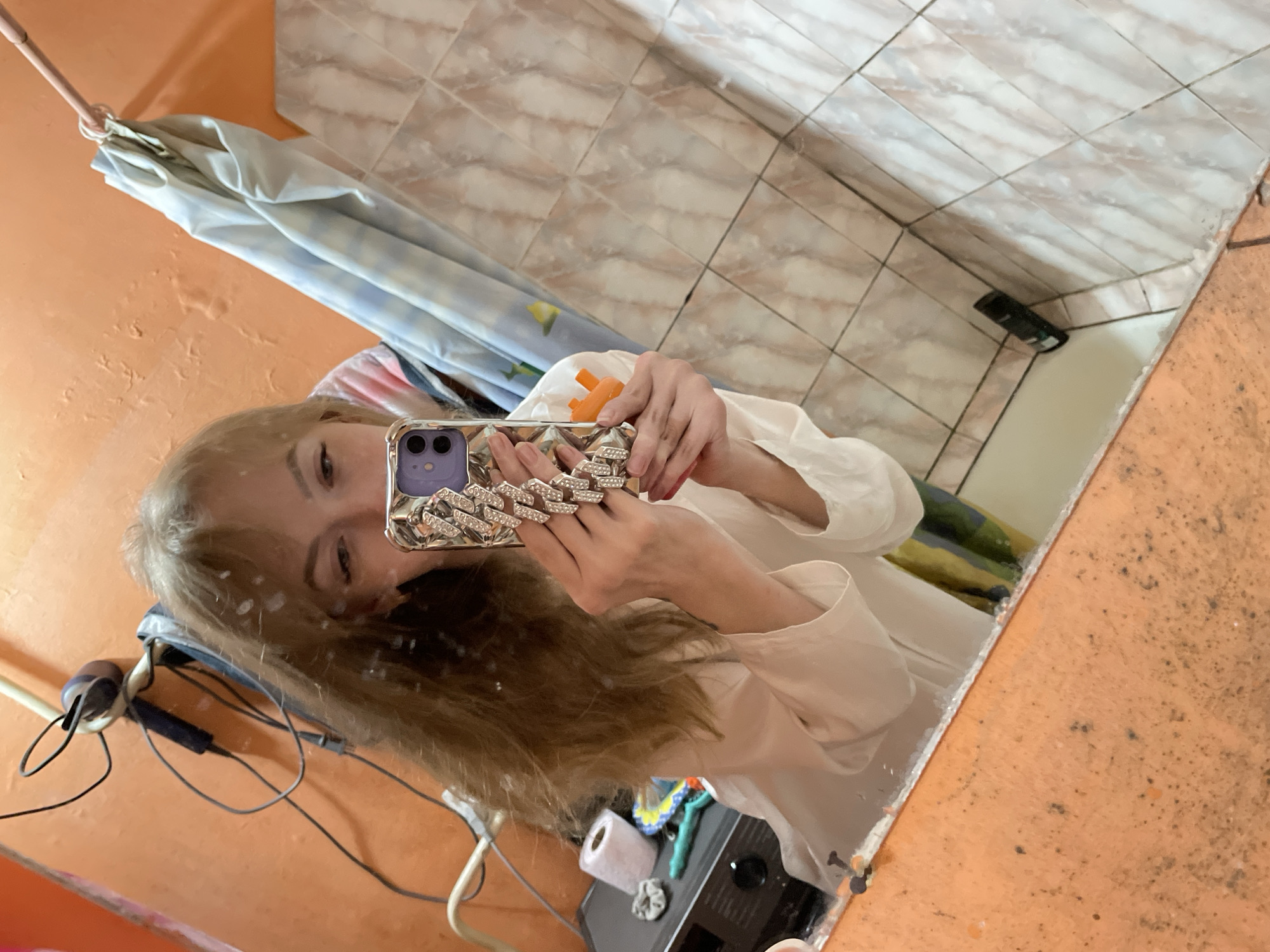 A young adult female with light brown hair is taking a selfie in a bathroom mirror. She is holding an iPhone in a diamond-encrusted case. Her reflection shows a shower curtain, tiled walls, a hair dryer, and bathroom accessories. The wall is painted orange. The location is possibly in Perm, Russia.