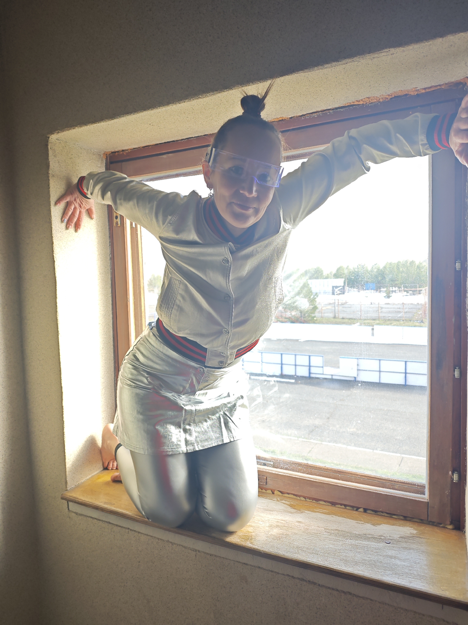 A woman in a silver outfit and clear safety glasses is crouched in a window frame. She is wearing a silver bomber jacket with a red and black striped collar and cuffs, a matching silver skirt, and silver leggings. Her hair is pulled back in a bun. She is holding onto the sides of the window frame with her hands. The window looks out onto a street and a fenced-in area. The location is Markovskii, Russia.