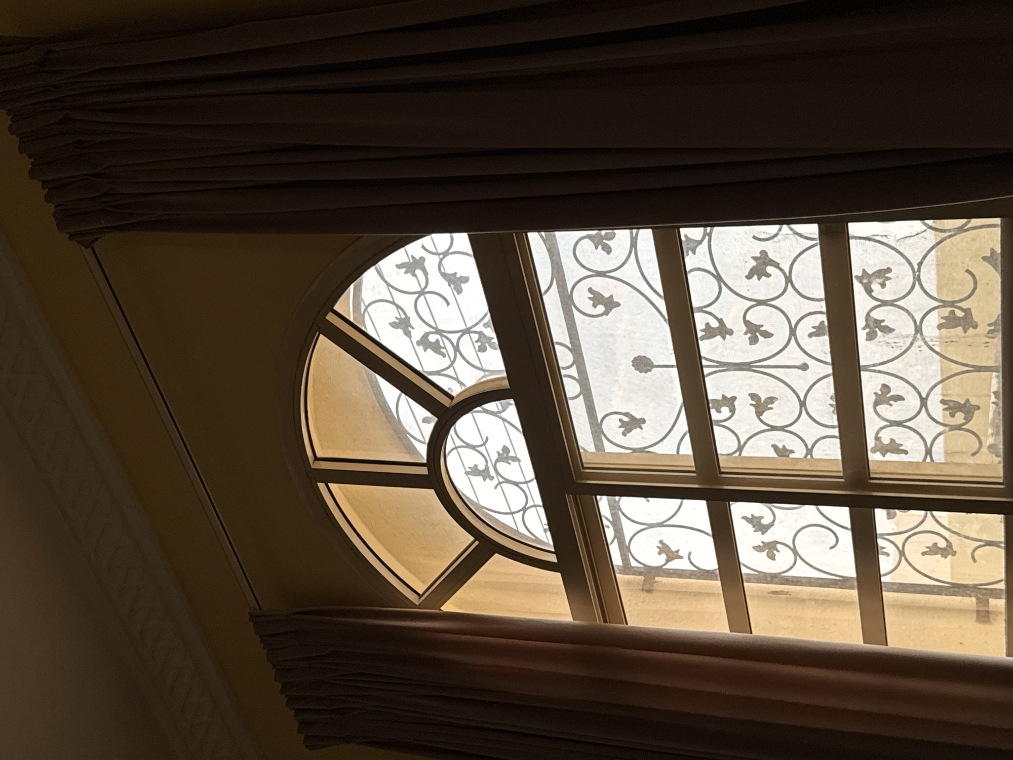 The image shows an arched window with decorative metal grills, partially obscured by brown curtains on either side. The window panes are illuminated by natural light, casting a warm glow on the yellow wall behind. The metal grills feature intricate scrollwork and leaf-like motifs. The scene appears to be an interior shot, likely in a residential or hospitality setting, given the architectural style and the presence of curtains. Without further context, it is impossible to definitively place this image in Al Buraimi Province, Oman, or any specific location.