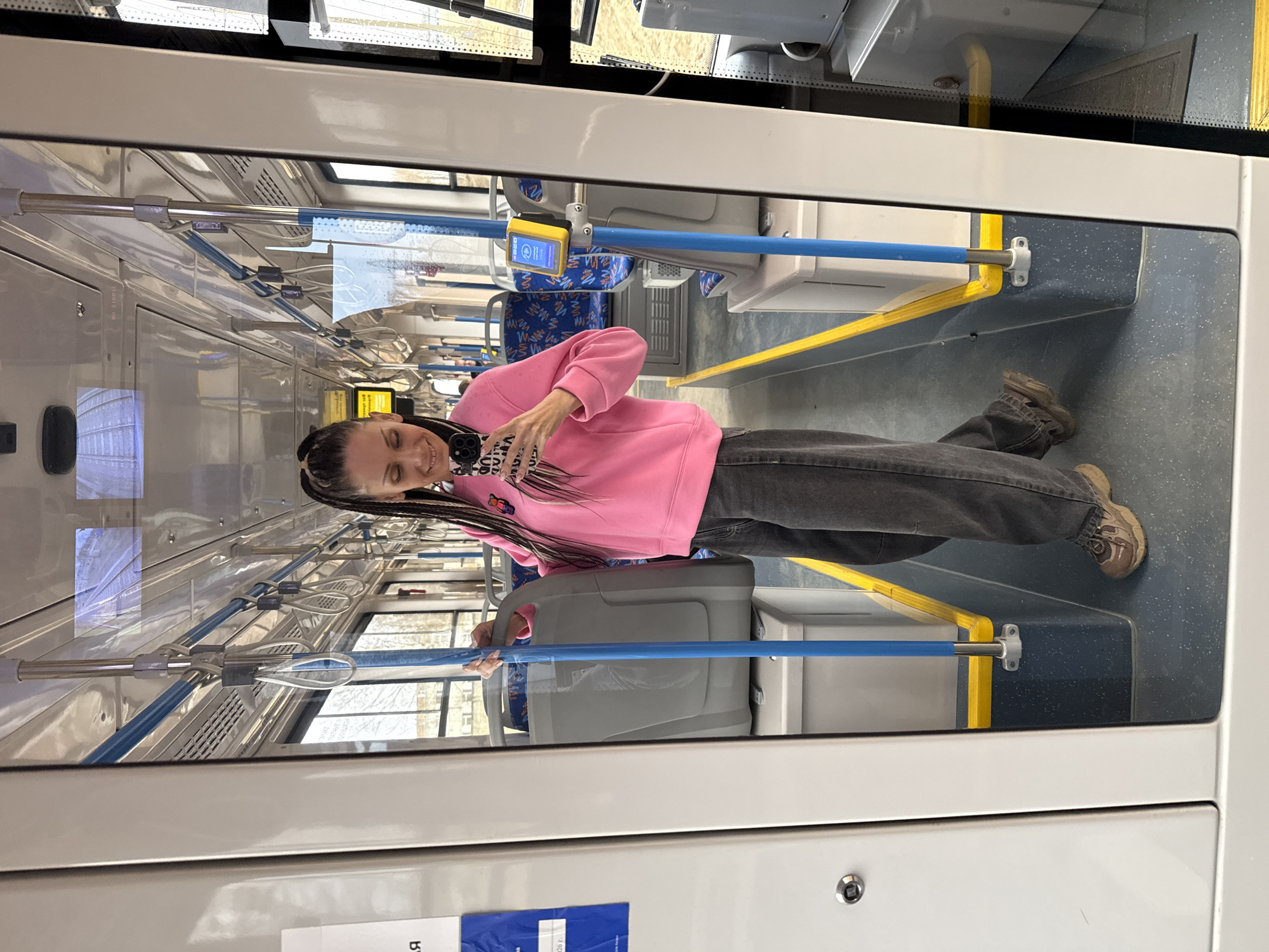 The image shows a woman standing inside a public transportation vehicle, possibly a tram or bus, which is the location. The woman has long, braided hair and is wearing a pink sweatshirt and gray jeans. She is holding a smartphone in her hand, possibly taking a selfie. The interior of the vehicle is equipped with seats, handrails, and electronic payment terminals. The city is Unknown, Unknown.
