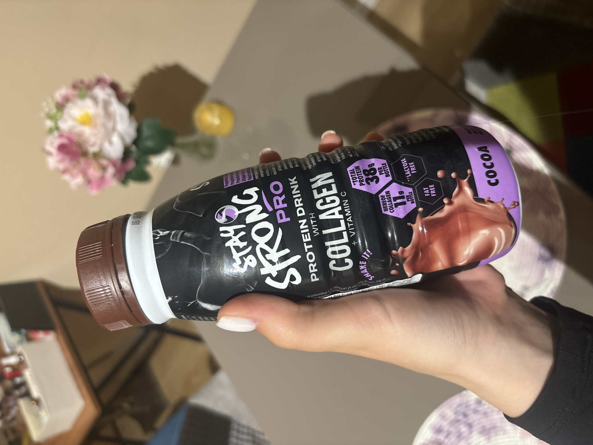 A fair-skinned person holds a bottle of "Stay Strong Pro" protein drink, cocoa flavor, in their right hand. The drink is black and purple, with text advertising its collagen and vitamin C content, as well as its protein and fat-free status. The background is out of focus, but shows a blurred floral arrangement and other furniture. The location is possibly indoors, though there's no information about the city of Sofia, Bulgaria.