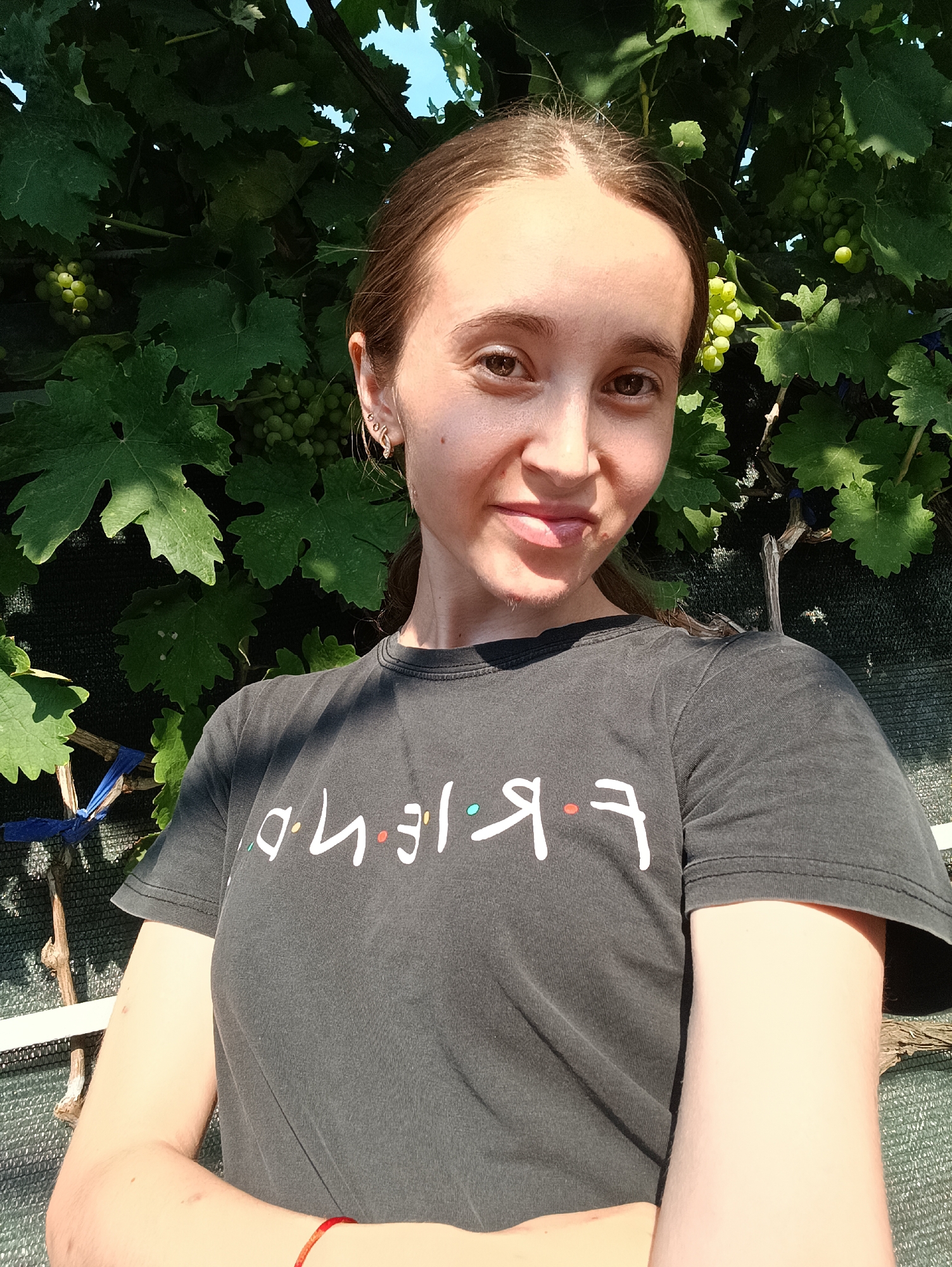 A young woman with long brown hair is taking a selfie outdoors. She is wearing a black t-shirt with the word "Friends" printed on it in white lettering with colorful dots between the letters. Her arms are crossed in front of her. She is standing in front of a dense green leafy vine with clusters of small green grapes. The background shows a black mesh fence. The image appears to have been taken during daylight hours. The location is Vypasne, Ukraine.