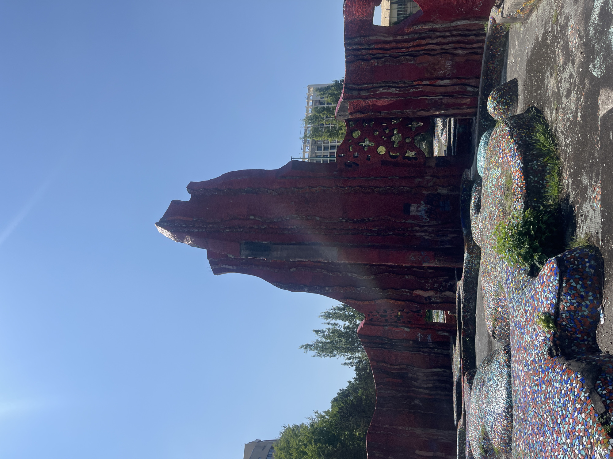 The image depicts the "Mosaic Playground" located in the city of Sochi, Russia. The playground features a large, abstract structure painted in vibrant red with vertical, textured lines, reminiscent of a rock formation or a stylized tree. The structure is adorned with colorful mosaic tiles in shades of red, blue, green, and white, particularly on the ground-level formations. The foreground is dominated by these mosaic-covered, undulating surfaces, interspersed with patches of dry earth and sparse greenery. The background reveals a clear blue sky, tall green trees, and modern buildings, including a multi-story structure under construction. The scene appears to be a public outdoor space, likely a park or recreational area, under bright daylight conditions. There are no people visible in the frame.