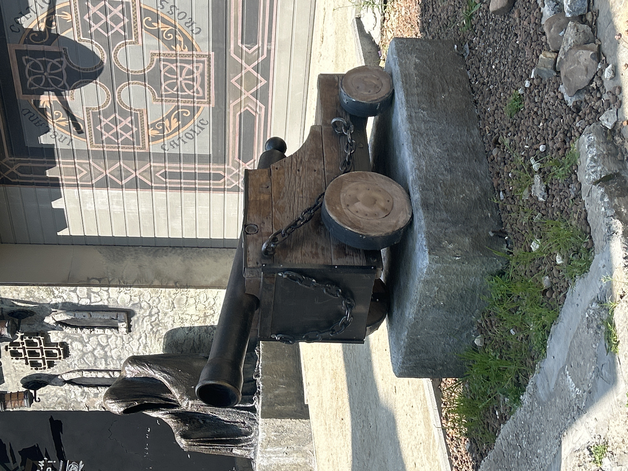 This is a full, eye-level, outdoor, daytime, eye-level medium shot of a historical cannon display. The cannon is made of wood and metal and sits on a stone pedestal in front of a building with a mural. The cannon is dark brown with a black barrel and wooden wheels. It is made to look old, with chains and visible screws. The pedestal is gray and rough. The building behind the cannon has a gray wooden facade with a mural depicting a Celtic cross and the words "CATHOLIC IRELAND CROSS". To the left of the cannon is a statue of a robed figure, possibly representing death. The background is blurry, but suggests a street scene. The ground is covered in gravel and small rocks. The location of this scene is the city of Perm, Russia.