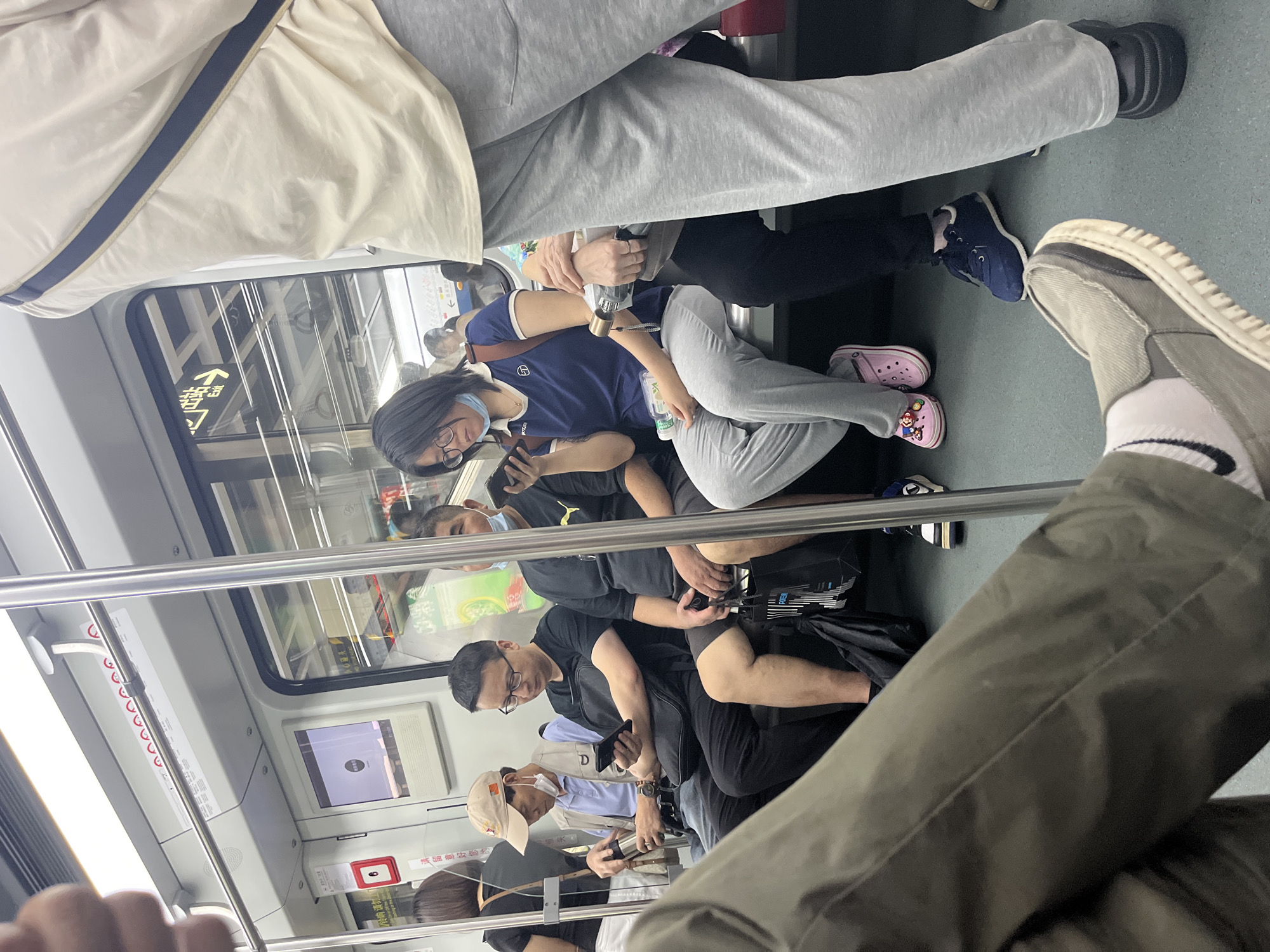 The image shows the interior of a subway train car with several passengers seated. In the foreground, a person's legs and feet are visible, wearing khaki pants and grey slip-on shoes with white soles. They are sitting and facing towards the left of the frame. Behind them, to the left, a man in a beige cap, blue polo shirt, and vest is looking down at a metal thermos. Next to him, a man in a black t-shirt and black pants is looking at his phone. To the right of him, a woman in a blue t-shirt and grey sweatpants is also looking at her phone, holding a plastic water bottle. She is wearing pink Crocs with character embellishments. Another person, mostly obscured by the foreground subject, is wearing grey sweatpants and a light-colored t-shirt with a blue stripe. Various bags and personal items are on the floor and seats. The environment is a typical subway train car with overhead lighting, metal poles, and windows showing a blurred view of the outside. Signs with Chinese characters are visible on the walls.