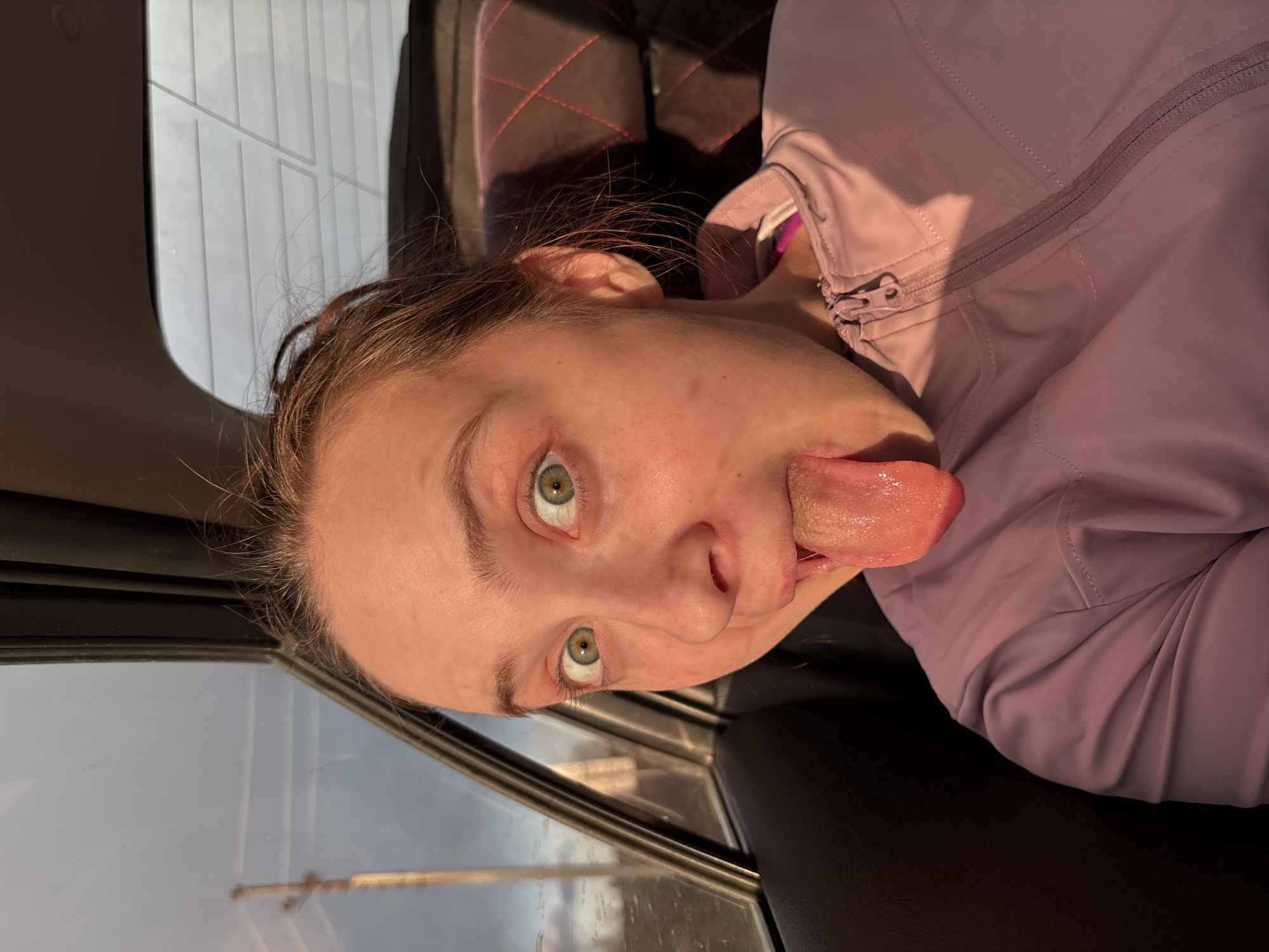 A close-up portrait shows a young individual, likely female, with light skin, light brown hair, and striking green or hazel eyes. The person is captured mid-expression, with their head tilted, eyes wide open, and tongue playfully sticking out to the side. They are wearing a light purple zip-up jacket.
The setting is inside a vehicle, indicated by the visible window frame and a dark, possibly leather, seat with a quilted pattern on the backrest. Through the window, an outdoor scene is visible, appearing somewhat blurred or distant, with hints of structures or poles against a light sky.
The lighting has a distinct warm, golden hue, suggesting the photo was taken during the late afternoon or early evening, often referred to as the "golden hour." The atmosphere of the image is candid and conveys a playful or humorous mood. There is no visible text in the image.