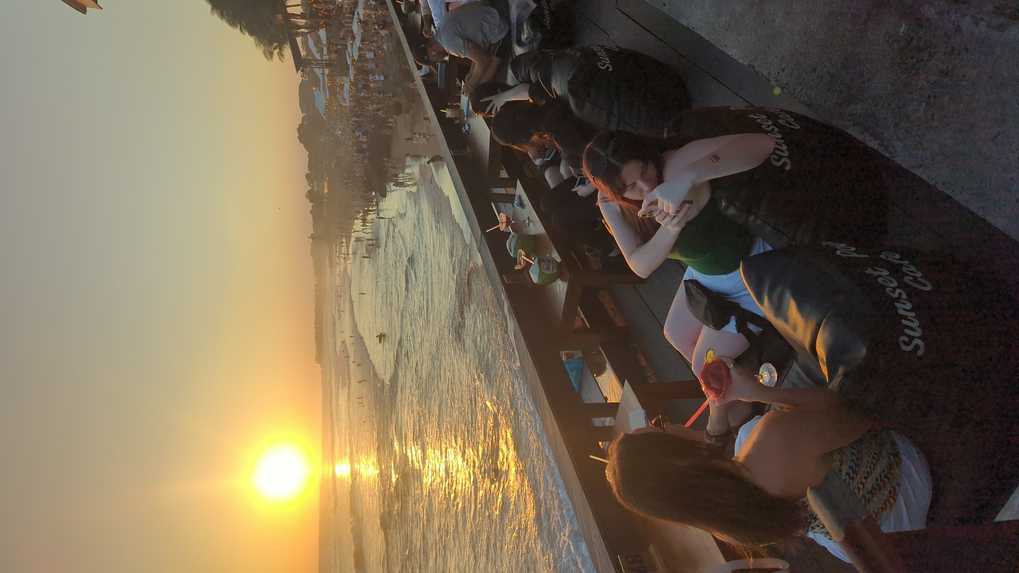 A scenic beachside view at sunset features patrons at an establishment identified as "Sunset Cafe" in North Kuta, Indonesia. The sun is setting over the ocean, creating a golden reflection on the water and a vibrant sky. A group of people sits at tables next to the ocean. One woman holds a cell phone, wearing a green top, while another sips a cocktail. Other patrons are also visible at the tables. The beach itself is populated with people enjoying the water, and buildings can be seen further away along the shoreline.