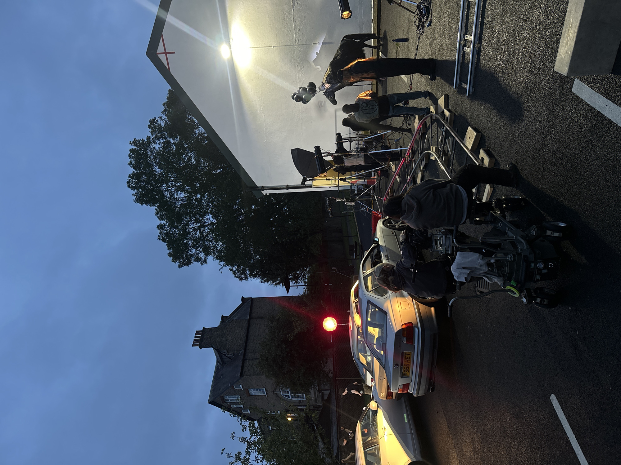 This is a medium shot of a film set at dusk on an overcast day, possibly in the Hackney or East London area. The scene is set in a parking lot or street with several cars, including a silver BMW and a white car. In the background, a large, old brick building with multiple windows and a prominent chimney stands to the left. To the right, a white building with a pitched roof and a red cross symbol is illuminated by a bright spotlight. In front of this white building, a dark horse stands tethered and is holding black balloons. A film crew is actively working around the horse, with lights, cameras, and dollies visible. Several people, dressed in casual clothing, are operating the equipment or observing the scene. The lighting suggests it is either early morning or late evening.