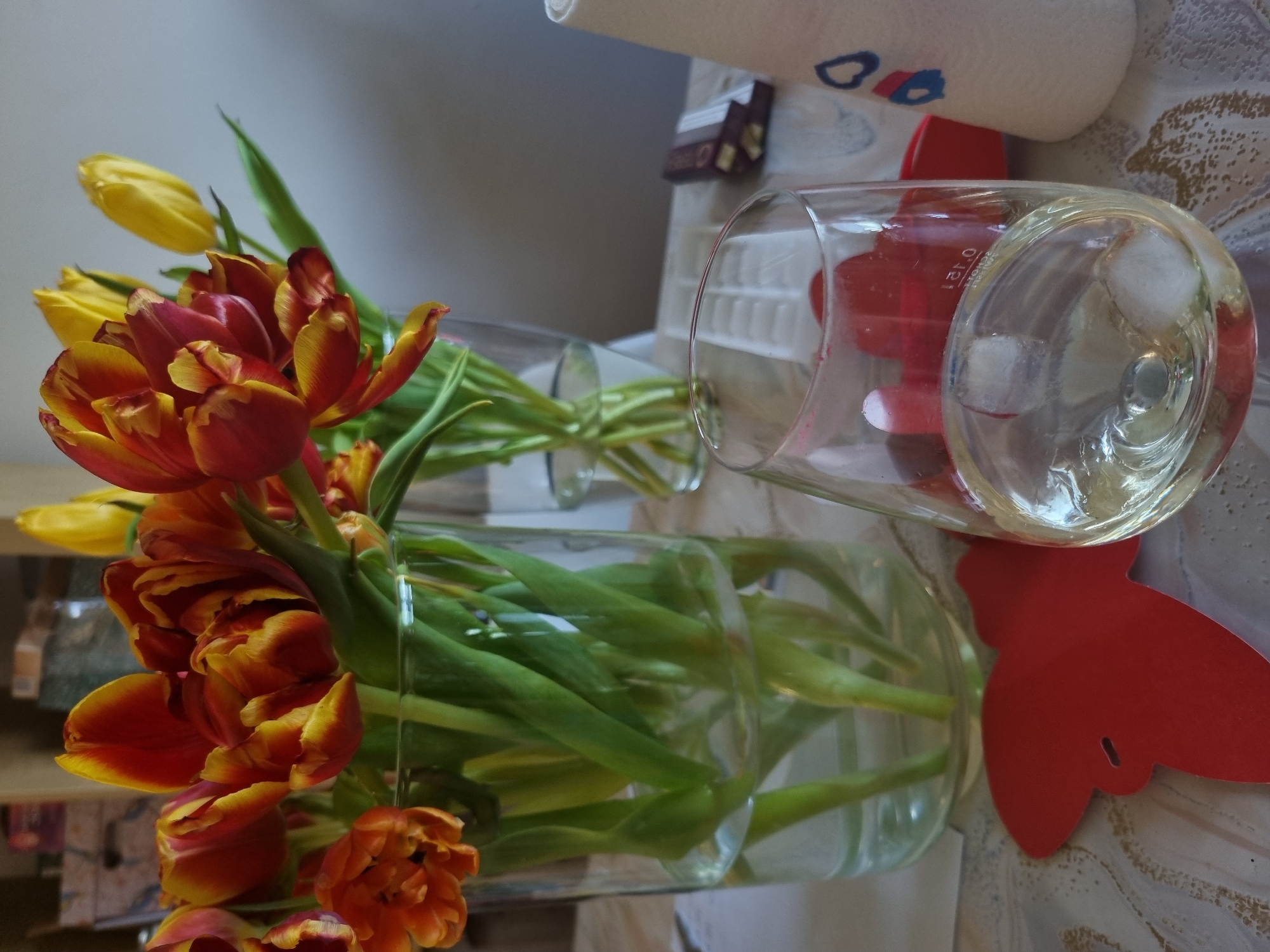 A still life image captures a vibrant arrangement of tulips placed on a patterned surface, likely a table, in what appears to be an indoor setting in Saarbrücken, Germany. The photograph is oriented vertically, showing the main subjects from a slightly elevated perspective.
On the left side of the frame, a generous bouquet of tulips fills two clear glass vases. The tulips display a striking mix of colors, predominantly fiery red and orange hues with prominent yellow edges, along with some pure yellow blossoms. Their vibrant green stems are visible through the transparent glass of the vases, submerged in water.
To the right, a clear glass, possibly a drinking glass or a beaker, rests on the same patterned surface. It contains a clear liquid and several ice cubes. A red marking, partially visible behind the glass and through its curve, indicates "0.12 l," suggesting a volume measurement. Next to this glass, a decorative red cutout, resembling a butterfly or a stylized floral shape, lies flat on the surface.
In the upper background, a roll of white paper towels or a thick napkin, adorned with blue and red heart-like patterns, is visible. Behind it, a small, dark-colored box with faint, unreadable text is partially obscured. The overall atmosphere conveyed by the bright flowers and light setting is cheerful and domestic.