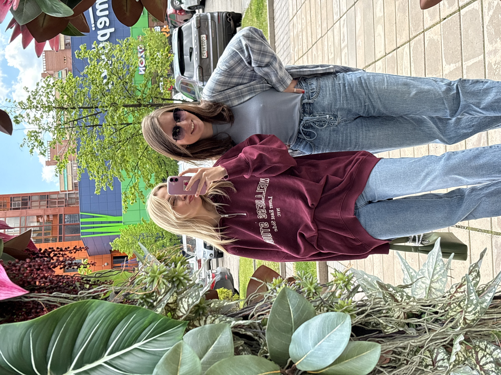 Two young women are standing on a paved surface outdoors, possibly a sidewalk. The woman on the left has blonde hair and is taking a selfie with a pink phone. She is wearing a maroon sweatshirt with white lettering and blue jeans. The woman on the right has brown hair, sunglasses, and is wearing a light blue top with a plaid jacket and blue jeans.
Behind them is a street scene with cars and buildings. One car has a license plate that appears to read "COOS BEST." The architecture in the background suggests an urban setting.
In the foreground is a decorative planter with large green leaves and various plants. The location is Krasnoyarsk, Russia, based on the website address displayed in the background.