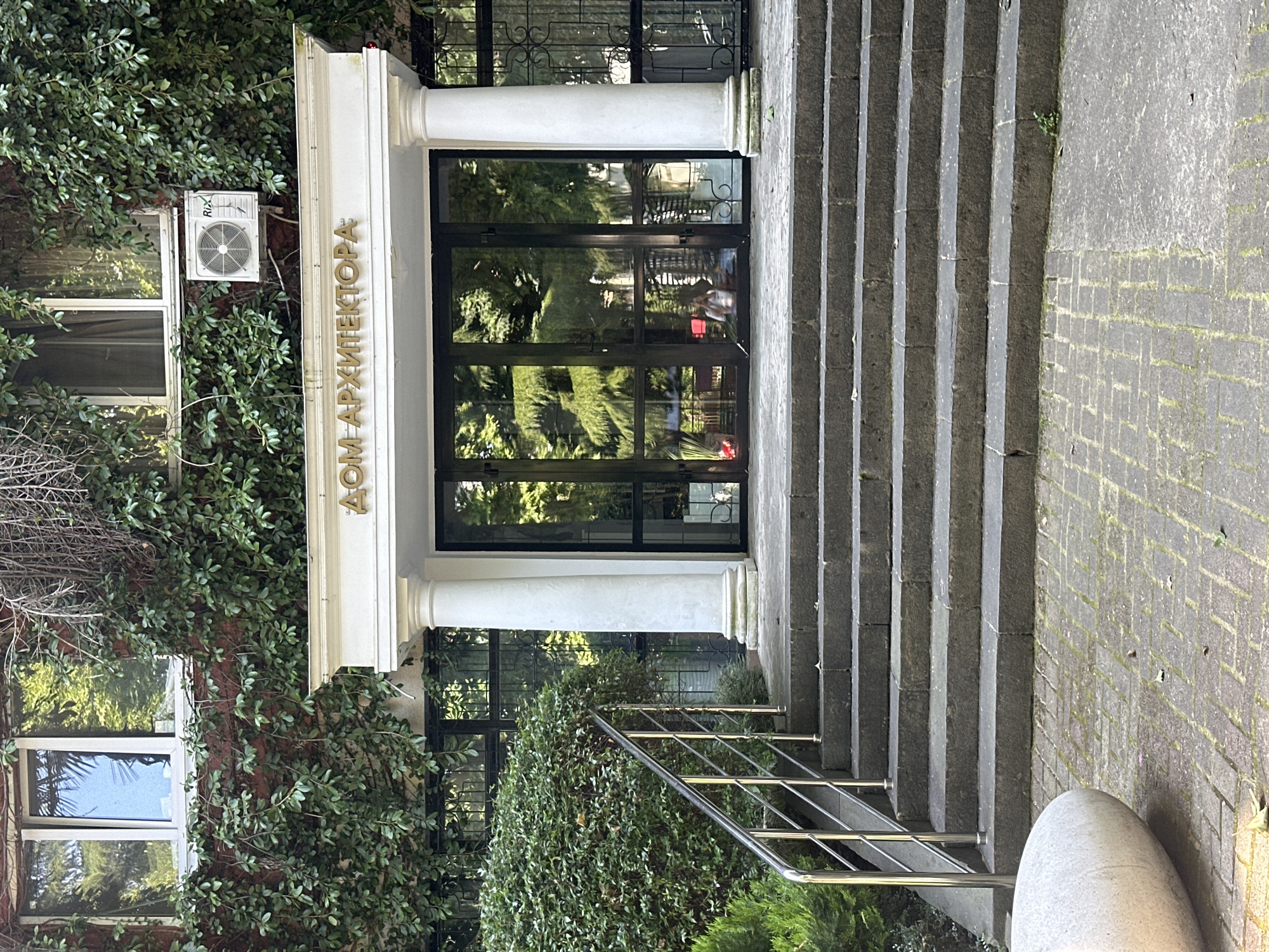 The image shows the entrance to a building covered in green ivy. The building has a white portico with two large columns, and above the glass doors, the words "ДОМ АРХИТЕКТОРА" are written in gold lettering. To the left of the entrance, there is a set of stone stairs with a silver railing, and a large smooth gray stone is positioned at the bottom of the stairs. The ground in front of the stairs is paved with cobblestones. A person is visible through the glass doors, sitting inside the building. The scene appears to be an exterior view of the Union of Architects of Russia building in Sochi, Russia.
