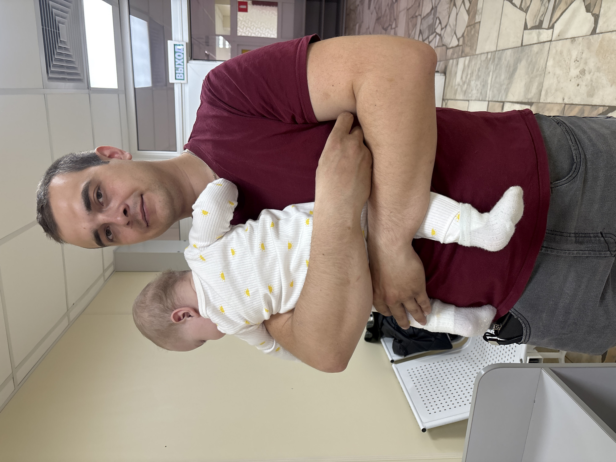 An image captures a man holding an infant in an indoor setting. The man, with short dark hair and a faint smile, wears a maroon t-shirt and grey jeans, looking directly at the camera. He cradles the baby in his arms, with the baby's head turned away, facing his chest. The infant is dressed in a white long-sleeved onesie adorned with small yellow sun or star patterns, and matching white socks.
The background suggests a public or commercial building, possibly a clinic or office. White ceiling tiles with an air vent are visible in the upper left, while a patterned tiled floor covers the right side. Behind the man, a door frame with glass panels is visible, and above it, a prominent green sign with white text reads "ВЫХОД" (VYKHOD), which is Russian for "EXIT." To the right, part of a white perforated plastic chair or stool and a grey container are visible. The overall lighting is bright and even, indicative of an interior environment during daytime. The scene suggests a calm moment between the man and baby, potentially in a waiting area in Klin, Russia.