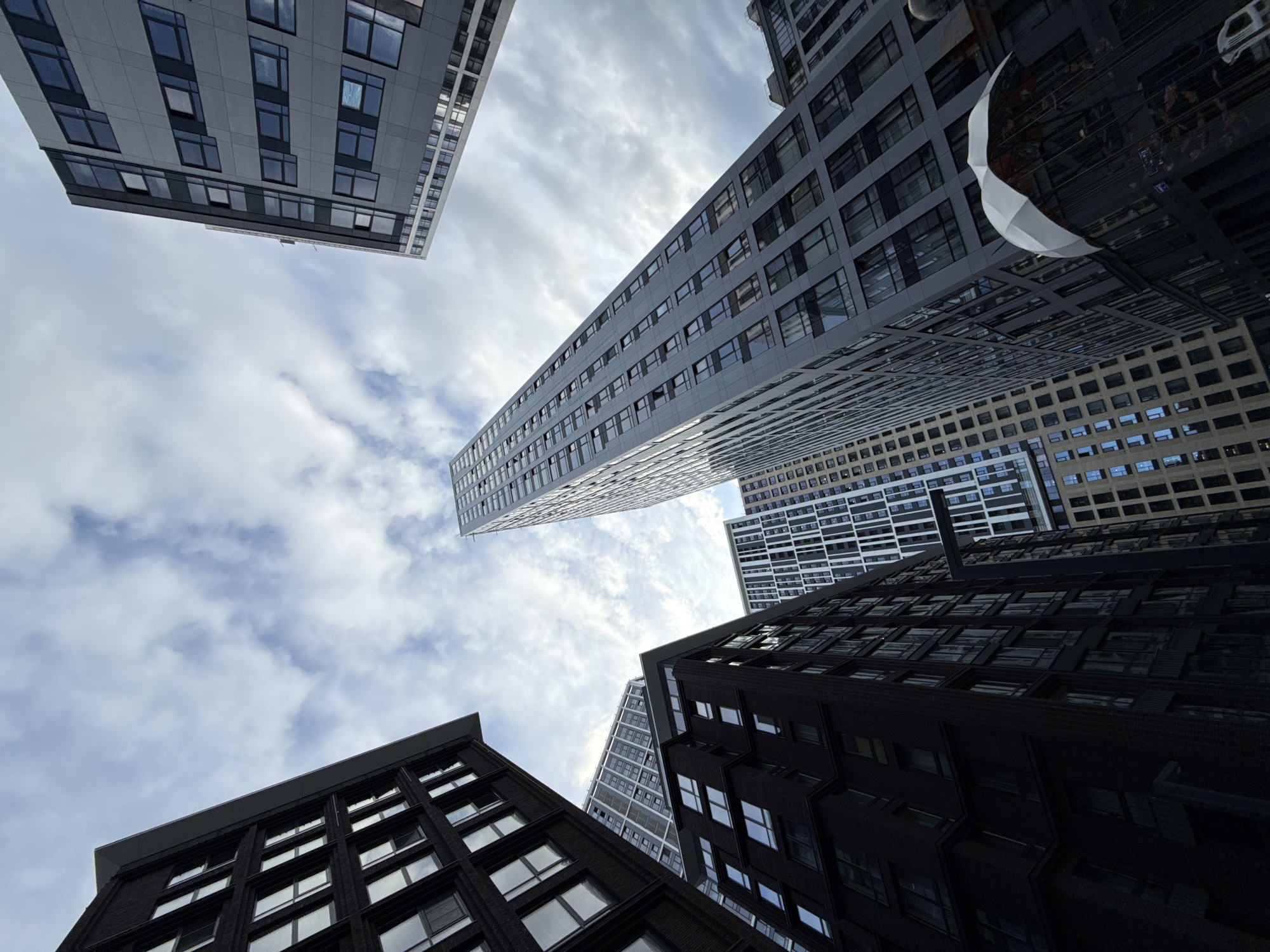 This is a low-angle shot looking up at a cluster of modern skyscrapers against a cloudy sky. The buildings are predominantly dark in color, with many windows reflecting the sky. One building in the foreground is clad in dark brick, with rows of regularly spaced windows. Another building, further back and to the left, is lighter gray and features a more grid-like facade with alternating dark and light rectangular panels. A very tall, slender building with a white, patterned facade is visible in the upper left of the frame. In the upper right, a portion of another building with a grid of small windows is visible. The clouds suggest it is daytime, possibly overcast or a partly cloudy day. The orientation of the buildings and the perspective create a sense of towering height and urban density. No people are clearly visible, and no text is discernable. The scene depicts a contemporary urban environment, likely a business or residential district.