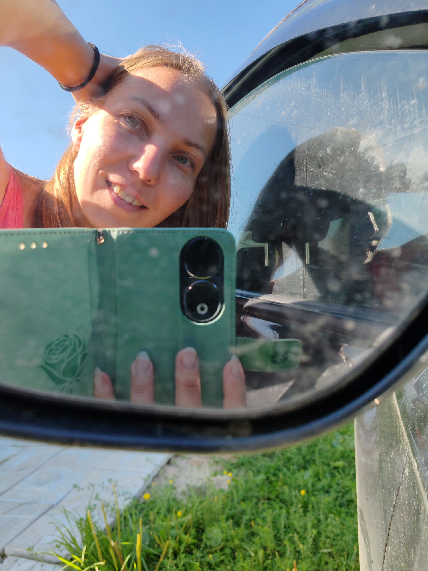 A young woman is taking a selfie in a side-view mirror of a vehicle. She has blonde hair and is wearing a pink top. She is smiling and holding her left hand up to her head. Her phone is visible in the mirror, which is a green case with a floral design. The background shows a clear blue sky, some grass, and a paved area. The setting appears to be outdoors in a rural or suburban area, likely in Чухлэм, Russia, given the context provided.