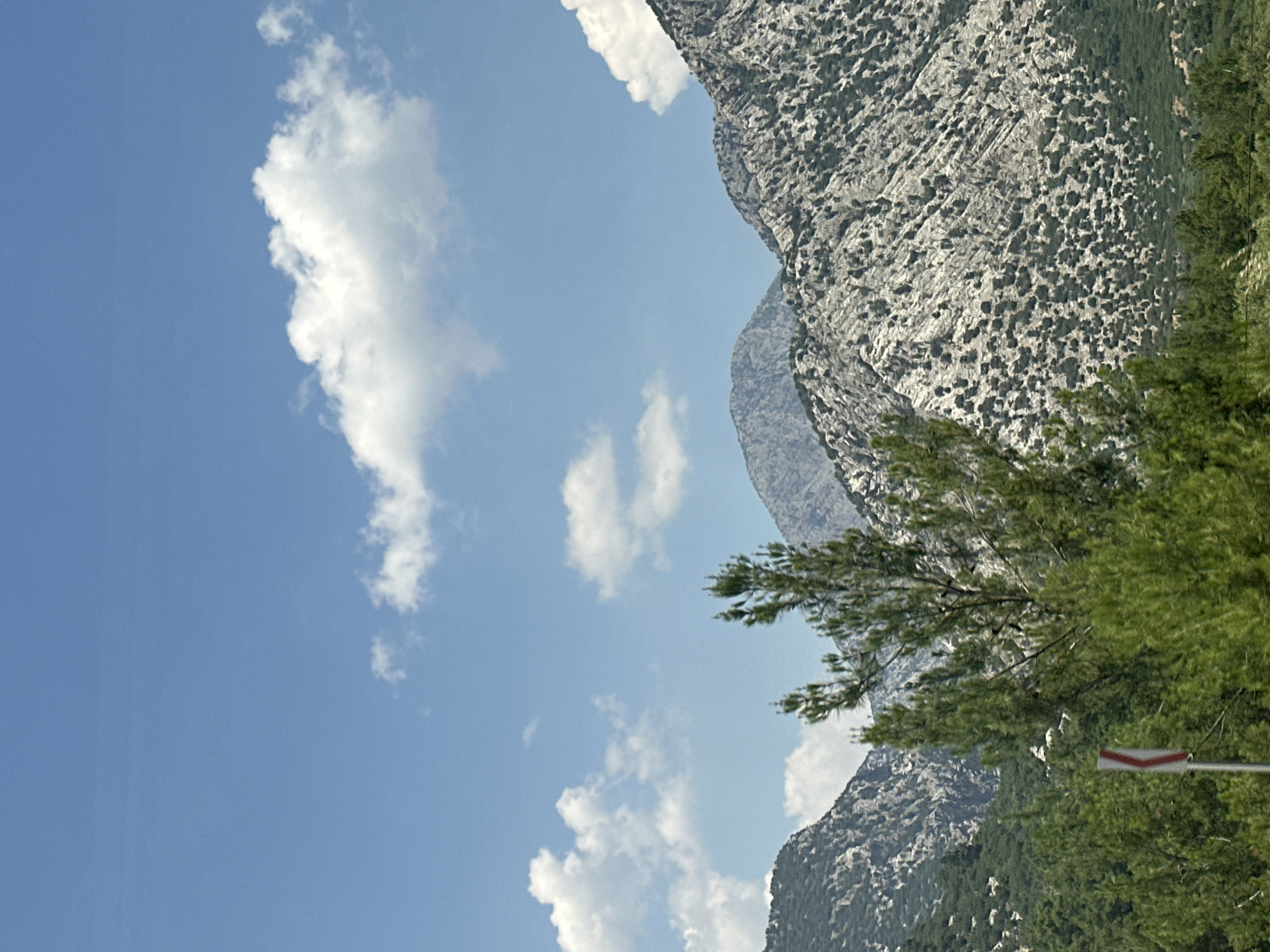 The image depicts a natural landscape with mountains and vegetation under a blue sky with scattered white clouds. In the foreground, there is lush green foliage, including a tall evergreen tree that partially obscures the view of the mountains. To the left, a red and white directional sign is visible amidst the trees. The mountains in the background are rugged and rocky, with patches of greenery on their slopes. The sky is clear and bright, suggesting a sunny day. The scene appears to be in a rural or mountainous area, possibly in or around Döşemealtı, Turkey, given the context. There are no people or animals visible in the image.