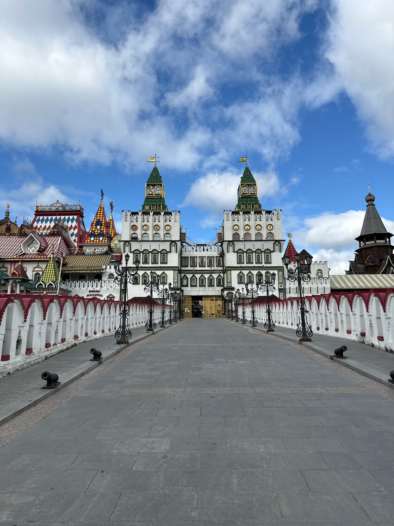 The image shows a wide, paved walkway leading towards a large, ornate building complex under a blue sky with white clouds. The complex features a central, white multi-story structure with green spired towers topped with flags. To the left and right of the central building are smaller, colorful structures with varied roof designs, including diamond patterns and pointed roofs. A white, red-capped wall with arched openings runs along the sides of the walkway, punctuated by ornate black lampposts and small black cannons. The walkway itself is paved with large, grey stone slabs. The Izmaylovo Kremlin in Moscow, Russia, is depicted in the image. No people are visible in the scene.