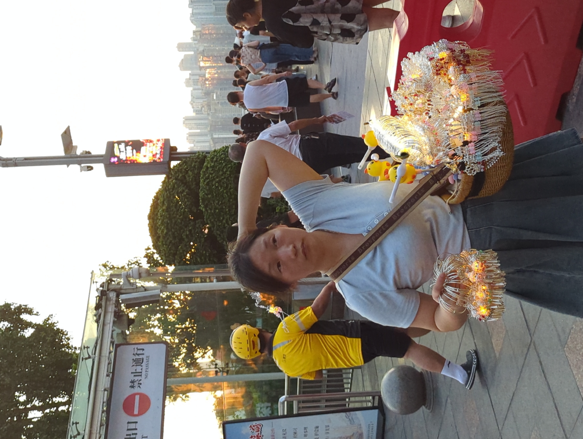 In Yuzhong District, China, a woman stands on a paved walkway, holding a woven basket filled with decorative trinkets. The trinkets appear to be small, silver charms and ornaments, some illuminated with small, colorful lights. She wears a light blue t-shirt and a gray pleated skirt. A strap with the text "BroSports" crosses her chest, supporting the basket. In her left hand, she holds a circular cluster of similar illuminated trinkets. Behind her, a man wearing a bright yellow shirt and helmet walks away from the camera. In the background, other pedestrians are visible, as well as buildings and trees. A sign with Chinese characters and "NO PASSAGE" is visible on the left. The sky is bright, suggesting it is daytime.