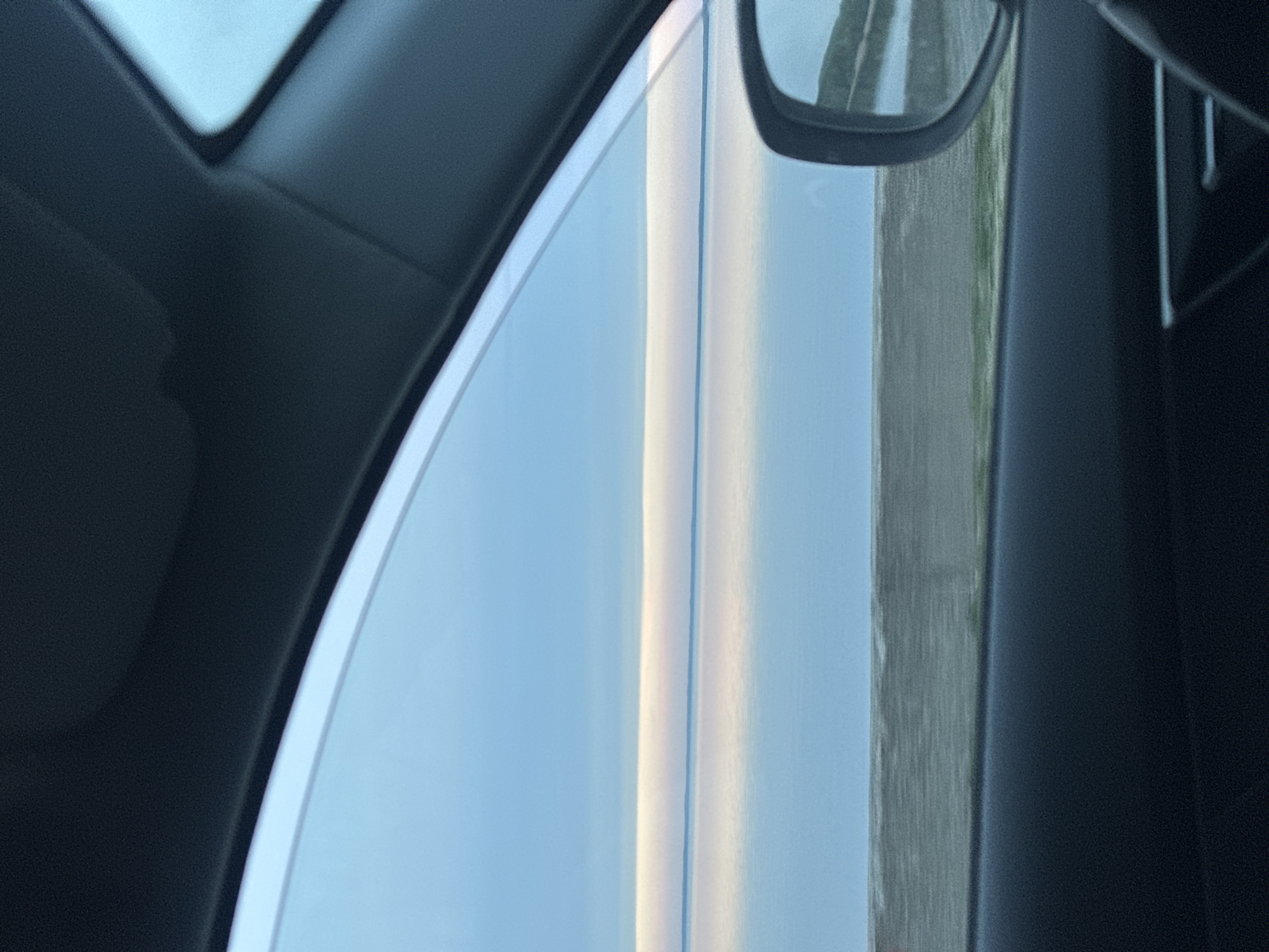 The image shows the view from inside a car, looking out of a side window and the side mirror. The scene outside the car includes what appears to be a landscape with a hint of greenery, likely a road or open field. The sky is visible in the window reflection. The interior of the car is visible, including the dashboard and door panel, which appear to be a dark color. There is no specific information in the image to indicate it is located in Chaikovsky, Russia. There are no people visible in the frame.
