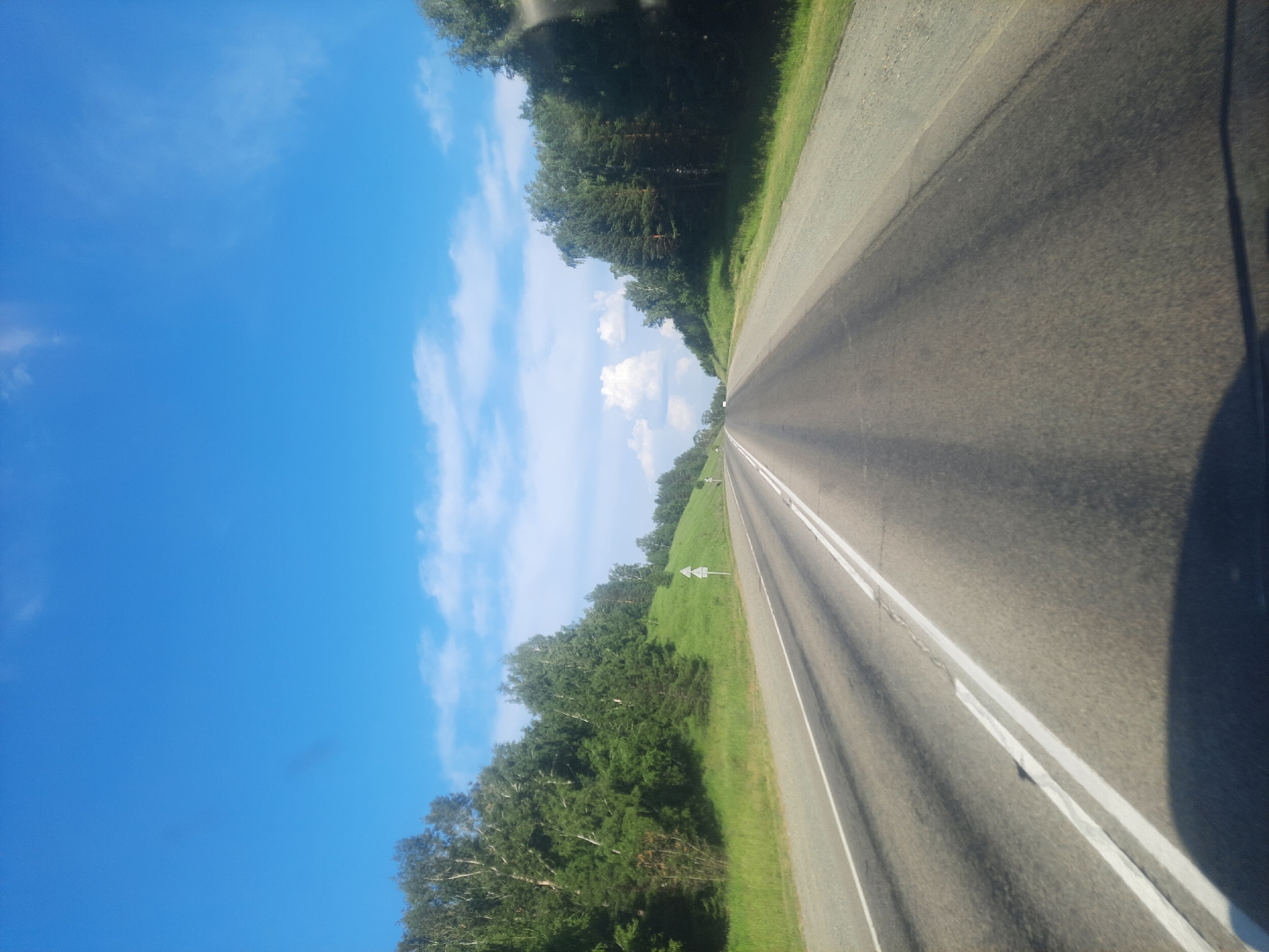 The media file captures a view from a moving vehicle on a paved road through a rural landscape in Баюновские Ключи, Russia. The scene is dominated by a bright blue sky with scattered white clouds, suggesting fair weather during daylight hours, likely late morning or afternoon. The road stretches forward, marked with double white lines, indicating a two-way highway. To the left of the road, a steep, grassy embankment is covered with lush green trees, primarily coniferous and some deciduous trees with white bark. A few white triangular road signs are visible on the embankment. To the right of the road, the view is partially obscured by the vehicle's shadow and a dark silhouette, possibly a rearview mirror or part of the vehicle's structure. The overall impression is a scenic drive through a natural environment on a sunny day. No people or specific events are depicted, only the landscape and the road.