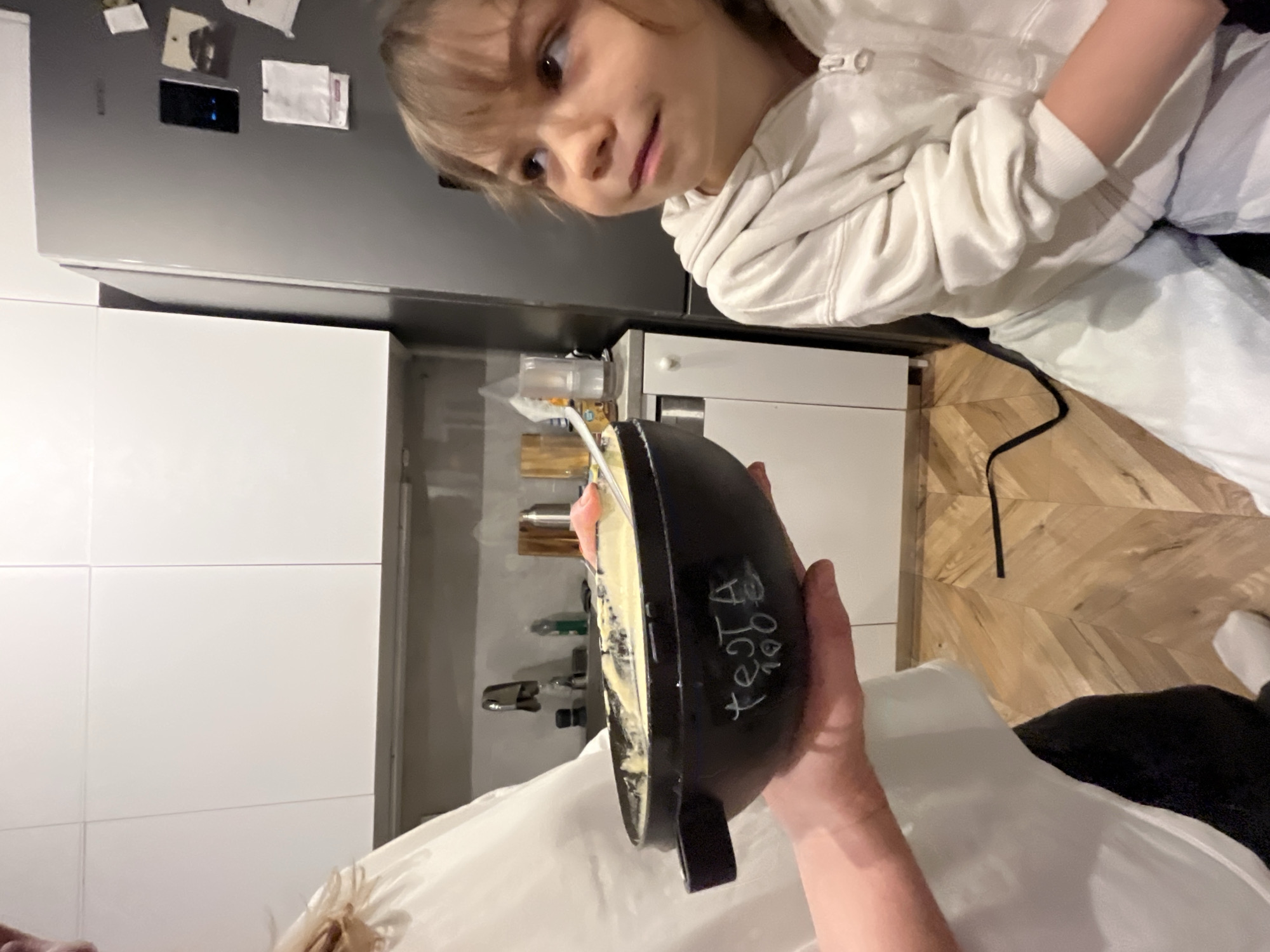 A person is holding a black frying pan containing some food, presumably containing an egg, in what appears to be a modern kitchen. A child is looking at the pan from above. The kitchen has white cabinets and a grey refrigerator with papers attached. The floor has wooden planks. The city of Budva, Montenegro cannot be determined from this image.