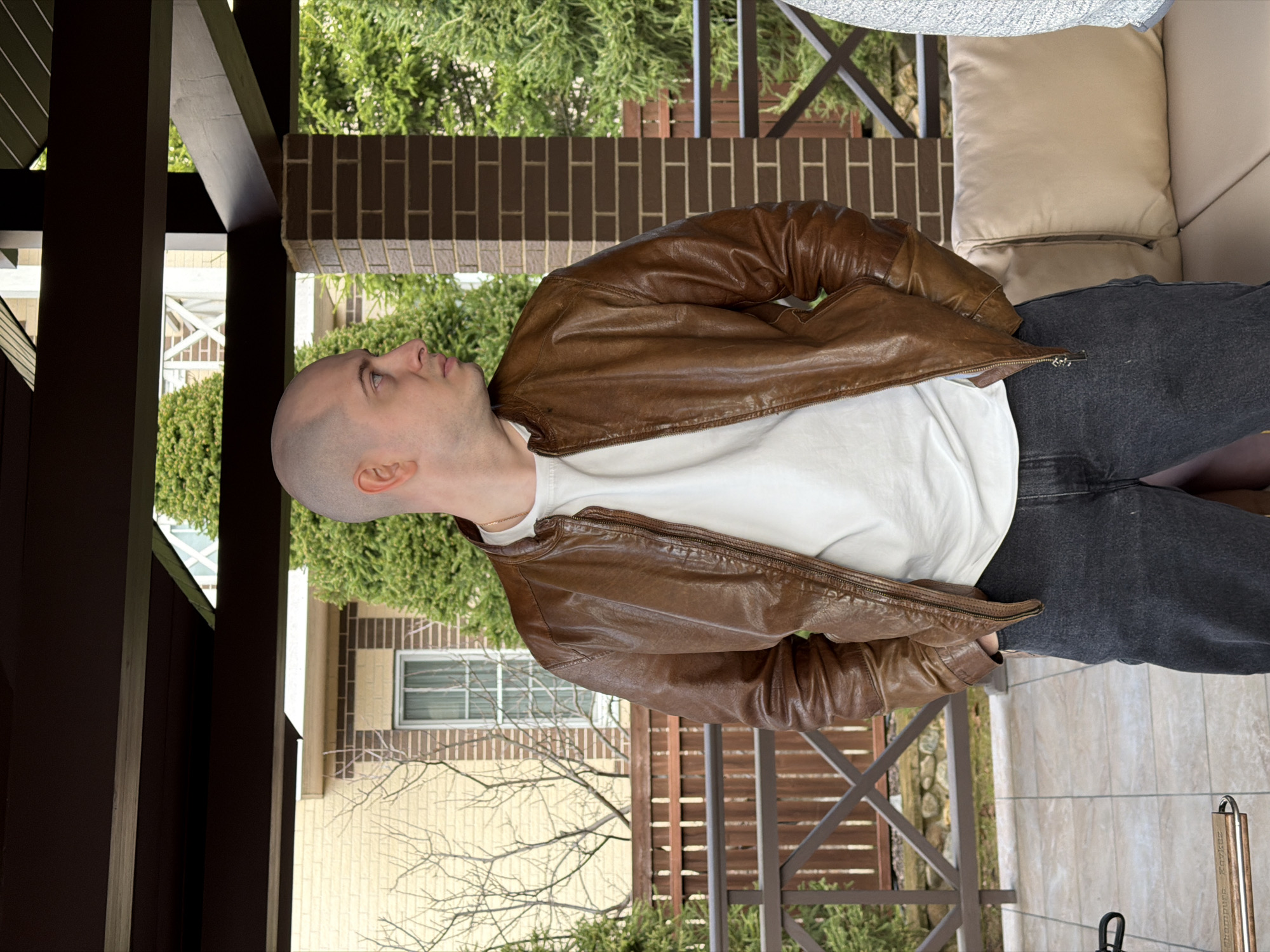 The image shows a bald man wearing a brown leather jacket over a white t-shirt and grey jeans. He is looking upwards and slightly to the left. The scene appears to be an outdoor setting, likely a porch or patio, with brick and wooden structures visible. Trees and foliage are present in the background, with a residential building partially visible. The location is specified as Mishutkino, Russia.
