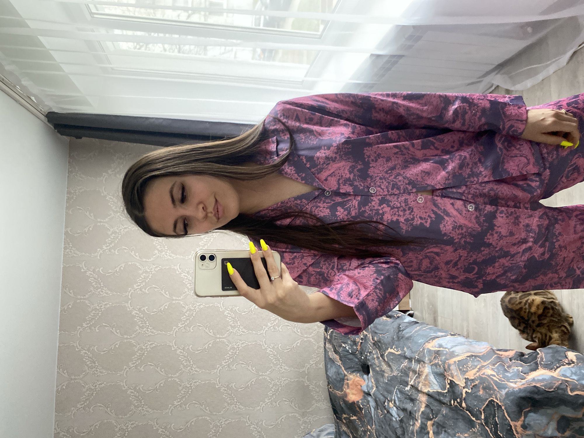 A young woman takes a selfie in a room with patterned wallpaper. She's wearing a pink and purple patterned pajama set. Her fingernails are painted bright yellow. She holds an iPhone in her right hand, and a black object is attached to the back of the phone. In the background, there's a window with blinds and a patterned bedspread. A Bengal cat is partially visible at the bottom right of the image. There's no information to confirm if the location is the city of Chaikovsky, Russia.
