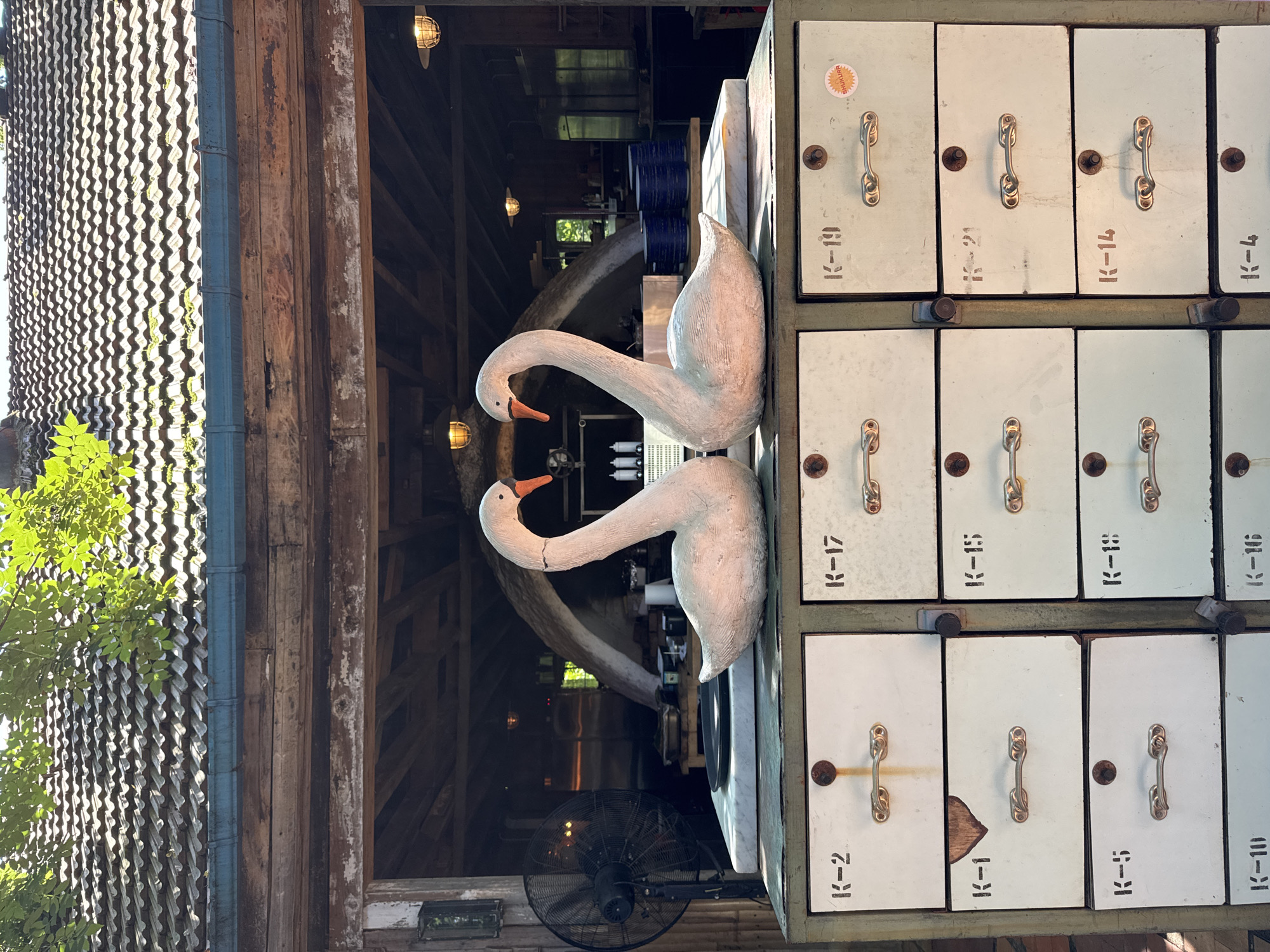 Two white swan sculptures, positioned to face each other with their necks forming a heart shape, are displayed on top of a multi-drawer cabinet. The cabinet has labels on each drawer, such as "K-2", "K-1", and "K-17". Behind the cabinet is an open-air structure with a tiled roof and wooden beams. The background shows a kitchen or restaurant interior with stainless steel appliances and shelves. A large black fan is visible to the left of the swans. The scene appears to be outdoors, possibly in a courtyard or dining area. The location is identified as Buknari, Georgia.