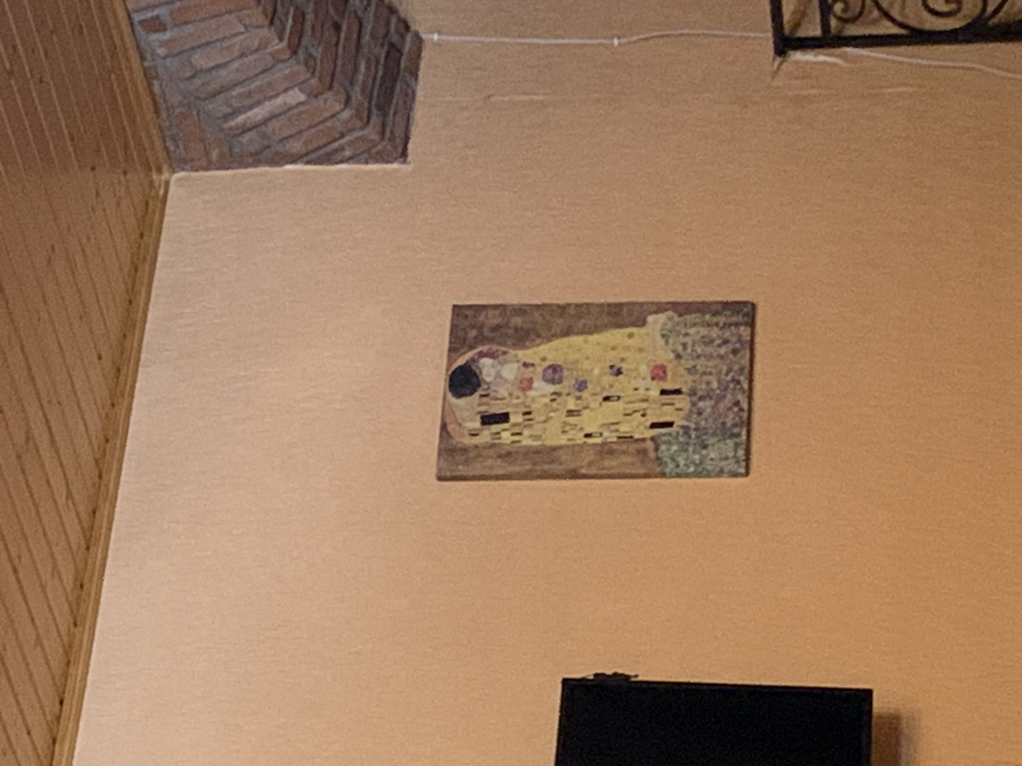 A framed print of Gustav Klimt's "The Kiss" is mounted on a textured, pale orange wall. The print depicts a couple embracing, with the man wearing a decorated robe and the woman in a floral dress, both appearing to kiss. The print is oriented vertically, with its bottom edge about waist-high on an average person. To the left of the print, a black television screen is partially visible, mounted on the same wall. The upper portion of the image shows a sloped wooden ceiling made of planks. To the right of the print, a brick archway is visible, with the bricks laid in a herringbone pattern. A white electrical cord runs vertically down the wall to the right of the print, secured by clips. In the lower right corner, a decorative black metal railing is partially visible. The lighting in the scene is dim, casting a warm glow on the walls and ceiling.