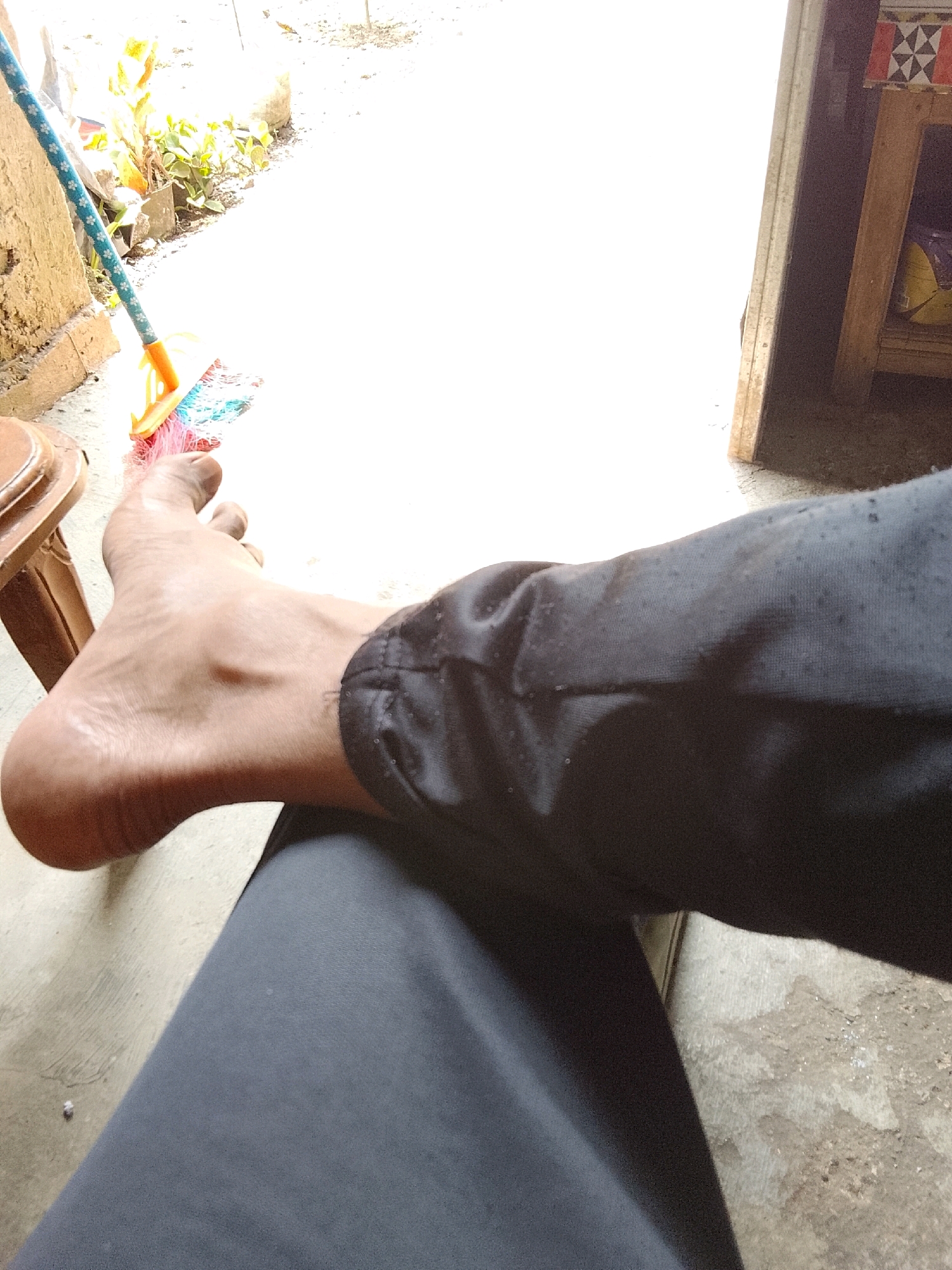 In Padang Dalom, Indonesia, a person's bare left foot is positioned on a light-colored concrete surface. The foot is pointing towards the right of the frame, with the heel and arch clearly visible. Dark fabric, likely pants, covers the lower leg, extending from the ankle down and across the lower portion of the image. To the left of the foot, a brown wooden stool is partially visible. Behind the stool and to the left of the foot, a broom with a blue patterned handle and a multi-colored brush (pink, yellow, and blue) rests against a rough, textured wall, possibly made of concrete or stone. The background on the right side of the image features a wooden structure with decorative geometric patterns, suggesting it is part of a building or furniture. The overall lighting indicates it is daytime, with bright light illuminating much of the scene, particularly the concrete ground.