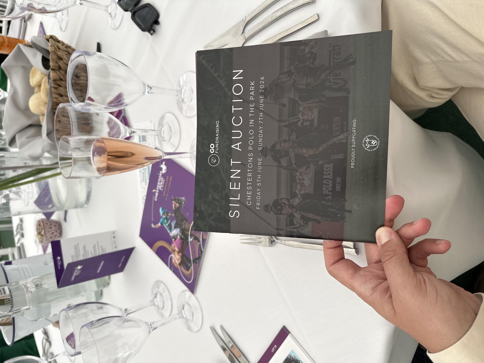 An eye-level close-up shot captures a person's left hand holding a dark grey brochure at a table setting, likely at a hospitality event associated with "Chestertons Polo in the Park" in Hurlingham Park, Fulham, London, United Kingdom.
The brochure, held by a light-skinned hand with visible fingernails and part of a light-colored sleeve, displays white text: "GO FUNDRAISING" with an accompanying logo, "SILENT AUCTION", "CHESTERTONS POLO IN THE PARK", and "FRIDAY 5TH JUNE - SUNDAY 7TH JUNE 2026". Below this text is a blurred image of polo players on horseback mid-match, with an audience visible in the background. At the bottom, "PROUDLY SUPPORTING:" is written above a circular logo, which appears to be for "THE SANDS END ASSOCIATED PROJECTS IN ACTION".
The table, covered with a white tablecloth, is set with various items. To the left, a silver fork is partially visible next to the brochure. Further left, a small purple card with "LOT 18" is partially visible. Behind the brochure, a pair of clear champagne flutes stand on the table, one containing a rose-colored liquid. Other clear glasses, possibly wine glasses, are also visible. A purple brochure with an image of a polo player and "CHESTERTONS POLO IN THE PARK" printed on it lies flat on the table. A folded purple tent card stands upright. In the mid-background, a woven basket holds several bread rolls, positioned behind a partially visible glass bottle. Another glass water bottle is visible further left, next to a white ice bucket. A small, patterned decorative bowl, possibly containing a condiment, sits on the table. To the right, a pair of black folded sunglasses rests on the white tablecloth next to a stack of silver knives. A person's leg, clad in light-colored trousers, is visible beneath the table on the right.