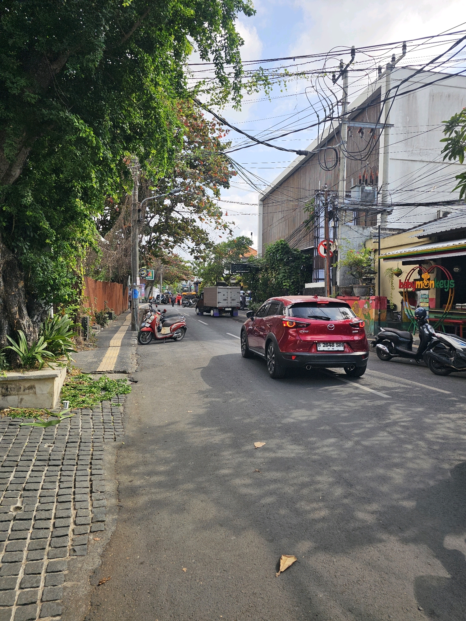 A red Mazda CX-3 SUV is driving on a street in Denpasar, Indonesia. The car is in the foreground, facing away from the camera. The street is paved with asphalt and has a white line marking the lane. To the left of the street is a sidewalk with a stone pathway. A red scooter is parked on the sidewalk. In the background, there are buildings with some trees and power lines. The sky is blue with some clouds. A truck is parked on the side of the road to the left of the red SUV. The overall scene is a typical urban street in a tropical climate.