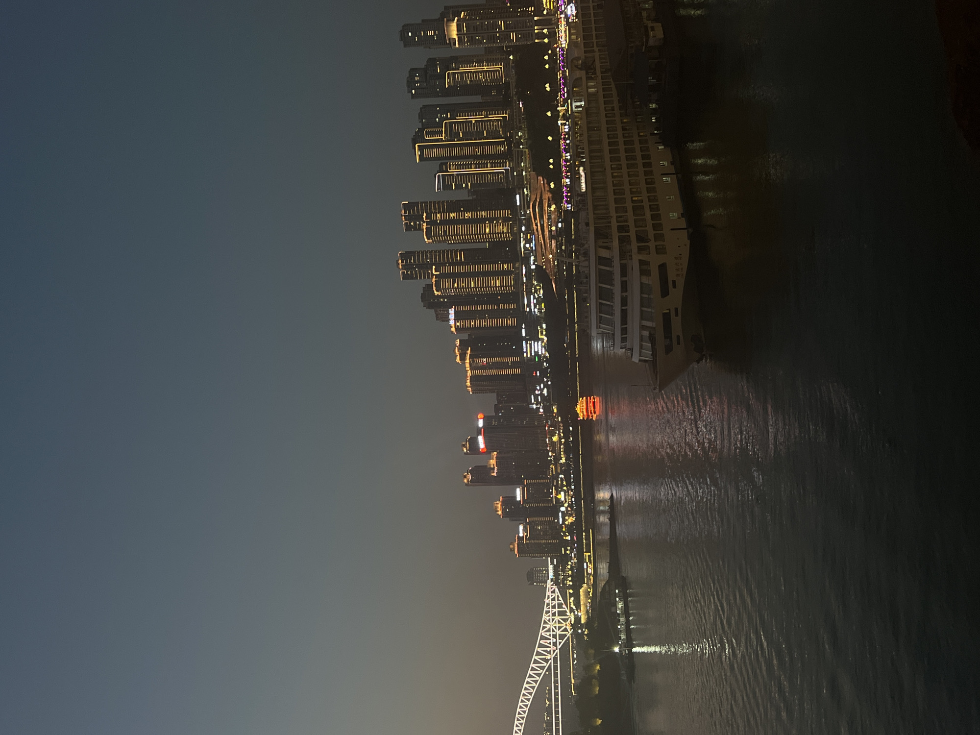 The image depicts a nighttime cityscape with a wide river in the foreground, likely the Yangtze River, adjacent to the Yuzhong District in China. A large, multi-decked cruise ship is docked on the right side of the river. The ship is illuminated, and its reflection is visible in the water. Across the river, a skyline of illuminated skyscrapers dominates the background. The buildings are lit with warm, golden lights, highlighting their vertical structures. A long, arched bridge, also illuminated with white lights, spans a portion of the river on the left side of the image. Several smaller boats are visible on the water, including one with a distinctive red, pagoda-like roof. The sky is dark and hazy, typical of an urban night. The overall scene is one of a bustling modern city at night, with emphasis on the illuminated architecture and the river as a central element.