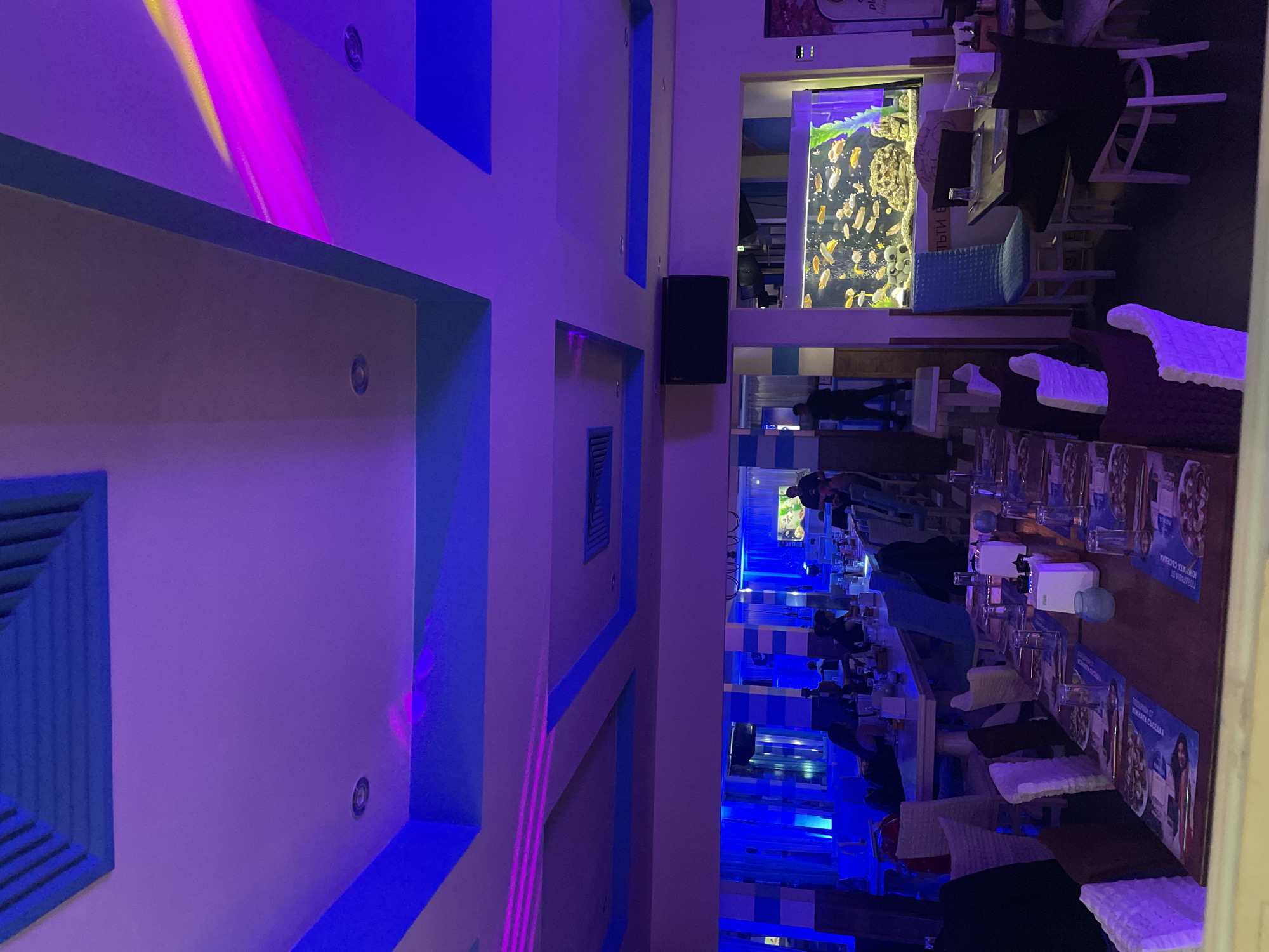 The image depicts the interior of a restaurant, likely in Plovdiv, Bulgaria, characterized by its distinctive blue-and-white decor. A prominent feature is an aquarium filled with fish, positioned between dining areas. Tables are set with glasses, menus, and napkins. Some patrons are visible, but their activities are not clearly discernible. The walls are adorned with geometric patterns and blue accents, contributing to the restaurant's aesthetic. The lighting scheme includes blue and purple hues, possibly from LED fixtures.
