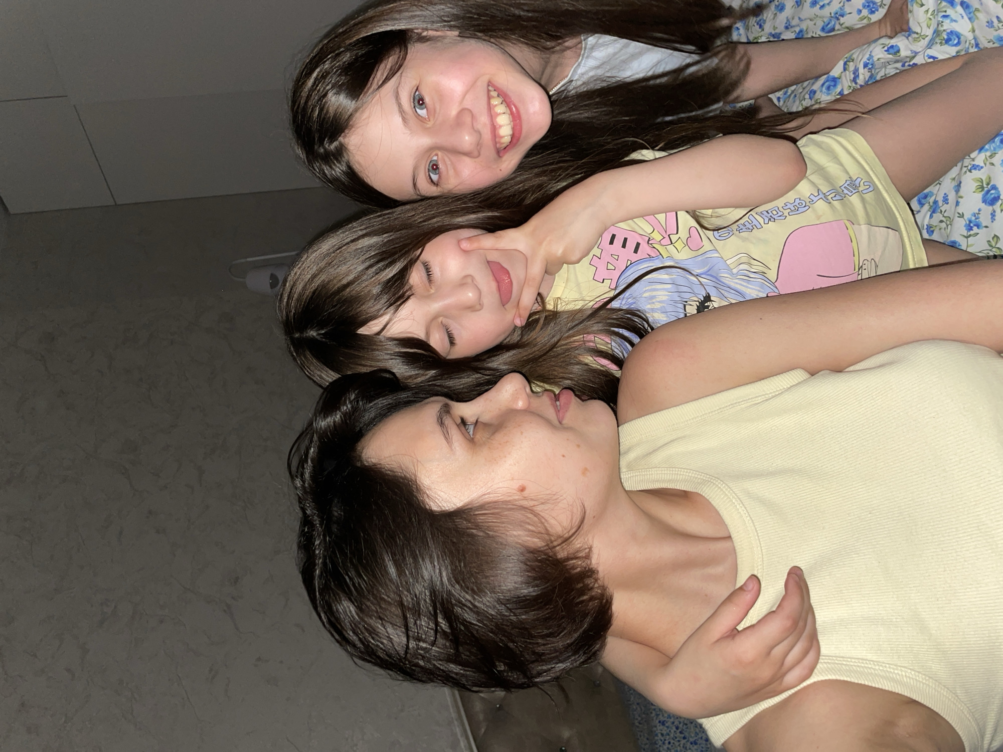 This image captures three females, likely two children and an adult, posed closely together in an indoor setting. The image is oriented vertically but appears to be taken from above, with the subjects lying down.
The uppermost subject is a young girl with long, dark hair, smiling broadly at the camera with her teeth showing. Her eyes are open, and she wears a light-colored top. Below her, another younger girl, also with long dark hair, playfully sticks out her tongue and makes a "peace sign" gesture near her face, with her eyes partially closed. She is wearing a light yellow t-shirt adorned with a colorful cartoon graphic that includes some Japanese-like characters.
At the bottom of the frame, an adult woman is visible in profile, looking towards the children. She has short to medium-length dark hair and freckles on her cheek. She is dressed in a light yellow ribbed tank top, and her arm is around one of the children, conveying a sense of warmth and affection.
The background shows a dark, textured surface to the left, possibly a floor or wall. On the right, a floral patterned fabric, featuring blue flowers on a white background, is visible, suggesting bedding or a blanket. The lighting is soft and subdued, creating an intimate and cozy atmosphere. The expressions and close proximity of the subjects convey a joyful, affectionate, and playful mood among family members or close companions. No specific outdoor or geographical identifiers for Izhevsk, Russia are visible within the image itself.