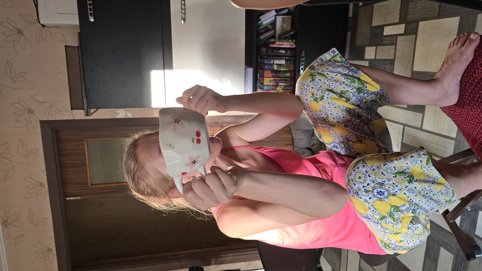 The image shows a woman sitting in a chair, holding a white cup with a floral and cherry pattern to her face. She is wearing a bright pink tank top and loose-fitting pants with a yellow and blue lemon pattern. The background reveals a room with floral wallpaper, a wooden door, and a black cabinet filled with books. The lighting suggests it is daytime, with sunlight streaming in from the right. The overall scene appears to be in a residential setting within Novohuivynske, Ukraine.
