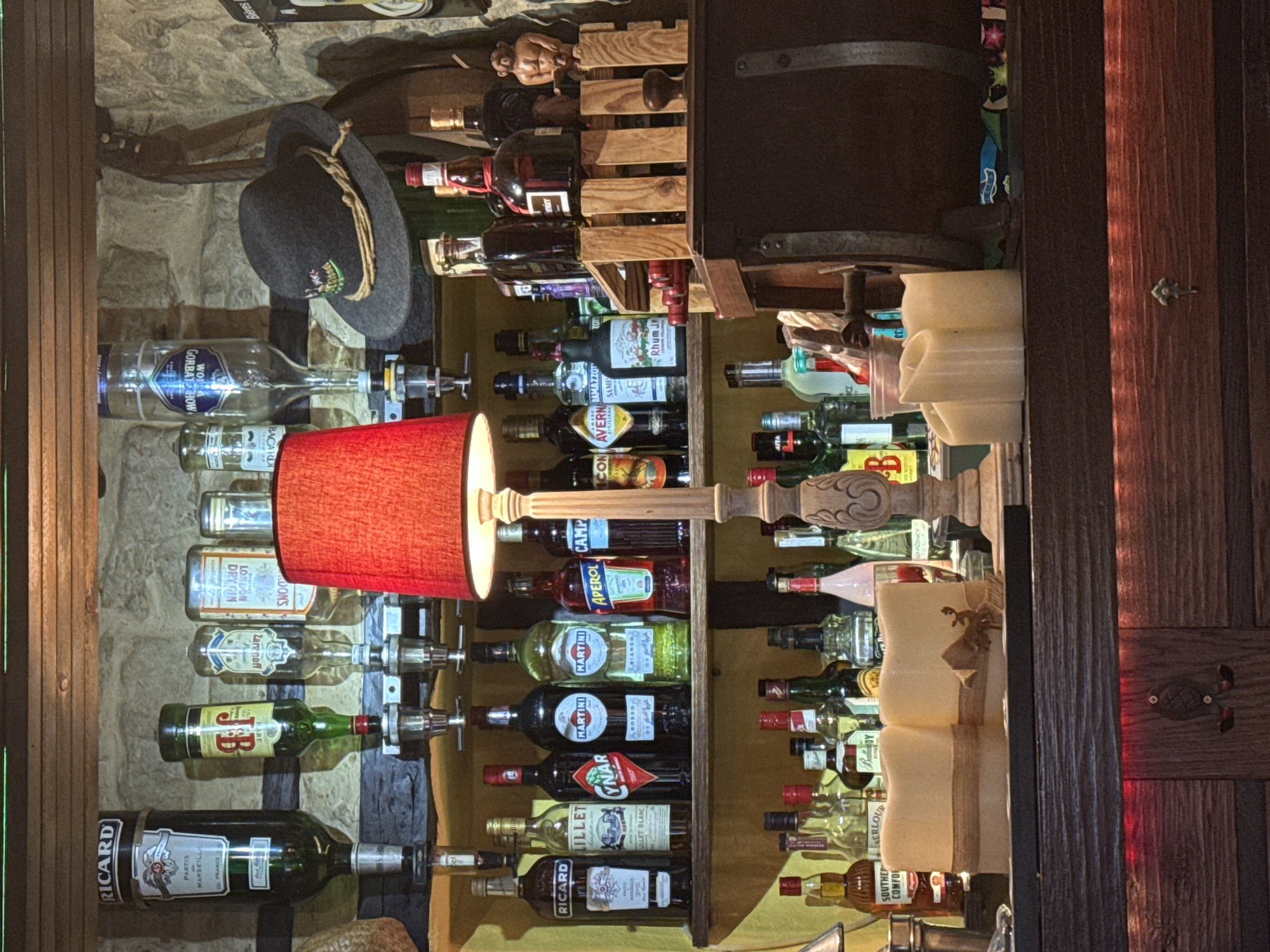 The image shows a vertical view of a bar display, likely in Junglinster, Luxembourg, given the presence of specific beverage brands commonly found there. The display features several shelves holding various liquor and wine bottles. A red lampshade illuminates part of the display. There is a wooden wine rack containing additional bottles. The display also includes decorative elements such as a traditional hat, a wooden sculpture of a figure, and stylized candle decorations. The background appears to be a stone wall, and a wooden cabinet or door is visible on the right side.