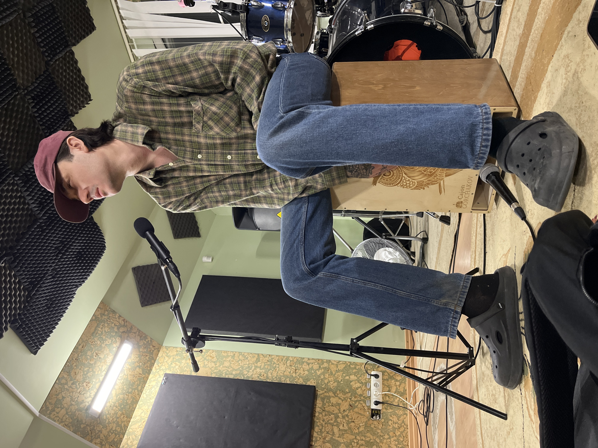 A person wearing a maroon baseball cap, a green and brown plaid long-sleeved shirt, blue jeans, and black Crocs is seated on a cajón in a room with soundproofing foam on the walls. A microphone on a stand is positioned near the cajón. A drum set is visible in the background. A white electric fan is behind the person. The location is unknown.
