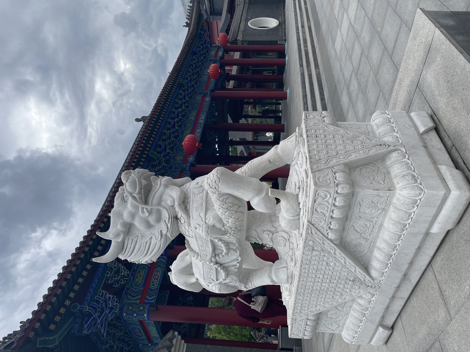 A white marble statue of a mythical creature, possibly a qilin or a dragon, stands prominently in the foreground of an outdoor courtyard. The statue is intricately carved and rests on a detailed pedestal. Behind the statue, a traditional East Asian style building with a multi-colored, ornate roof and red pillars is visible. Several red lanterns hang from the building's eaves. The sky is overcast and gray. A person in a dark red garment walks away from the camera towards the building, carrying a bag. The courtyard is paved with gray tiles, and steps lead up to the building. The overall scene suggests a historical or cultural site.