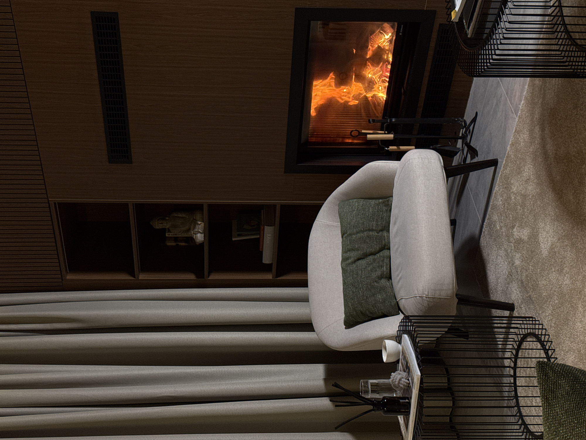 The image depicts an indoor scene, likely a living room, with a fireplace as the focal point. To the left, a light-colored armchair with a green pillow is placed near a side table. The side table holds a glass of water, a diffuser with reeds, and a small white bowl. Behind the armchair and side table, grey curtains are drawn. A bookshelf filled with various items, including a statue and books, is visible on the left wall. The fireplace on the right wall is lit, with flames and glowing embers visible. Next to the fireplace, a set of fireplace tools stands on the floor. A wooden wall paneling is present on the right side of the room. The floor is covered with a light-colored rug. The overall ambiance is warm and cozy. The location is identified as Bakovići, Montenegro.