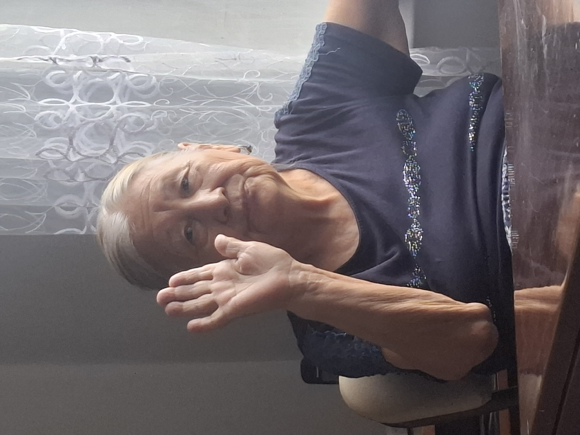 An elderly woman with graying hair is sitting at a table in front of a window with sheer white curtains. She is wearing a dark blue t-shirt with a decorative trim on the sleeves and a sequined design across the chest. The woman is raising her right hand, palm facing forward, in a waving gesture. Her left arm is resting on the armrest of a chair. The background shows a dark wall on the left and the window with curtains on the right. The scene appears to be indoors, possibly a home or a room in Almaty, Kazakhstan.