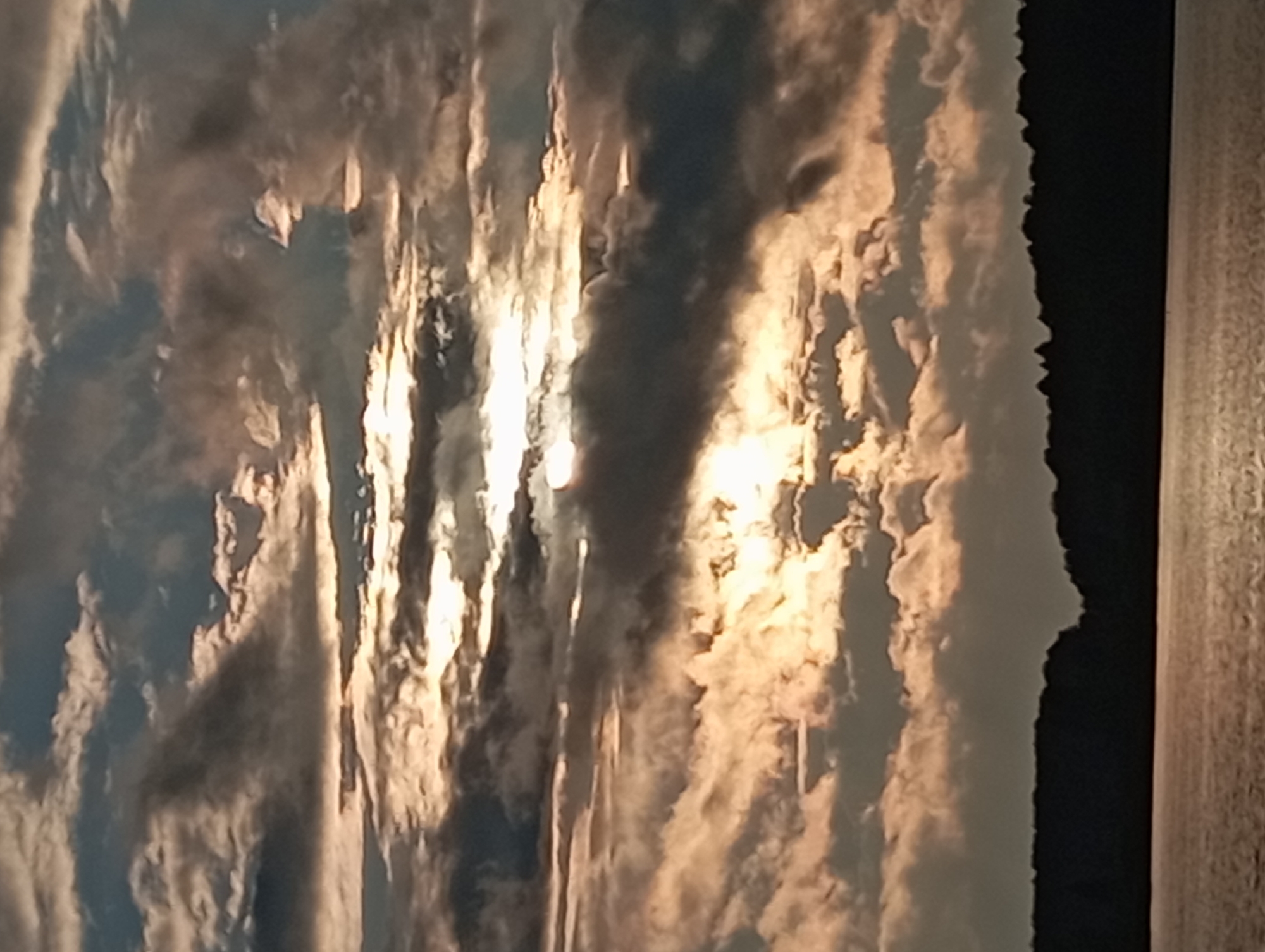 The image depicts a dramatic sunset over a body of water, likely a lake, in Mołowiste, Poland. The sky is filled with heavy, textured clouds, illuminated by the setting sun which peeks through in several areas, casting a warm, golden glow. Darker, silhouetted clouds create a stark contrast against the brighter ones. Below the sky is a dark, silhouetted treeline that forms the horizon. The water in the foreground reflects the muted colors of the sky, with gentle ripples visible on its surface. The scene captures a moody and atmospheric natural landscape.