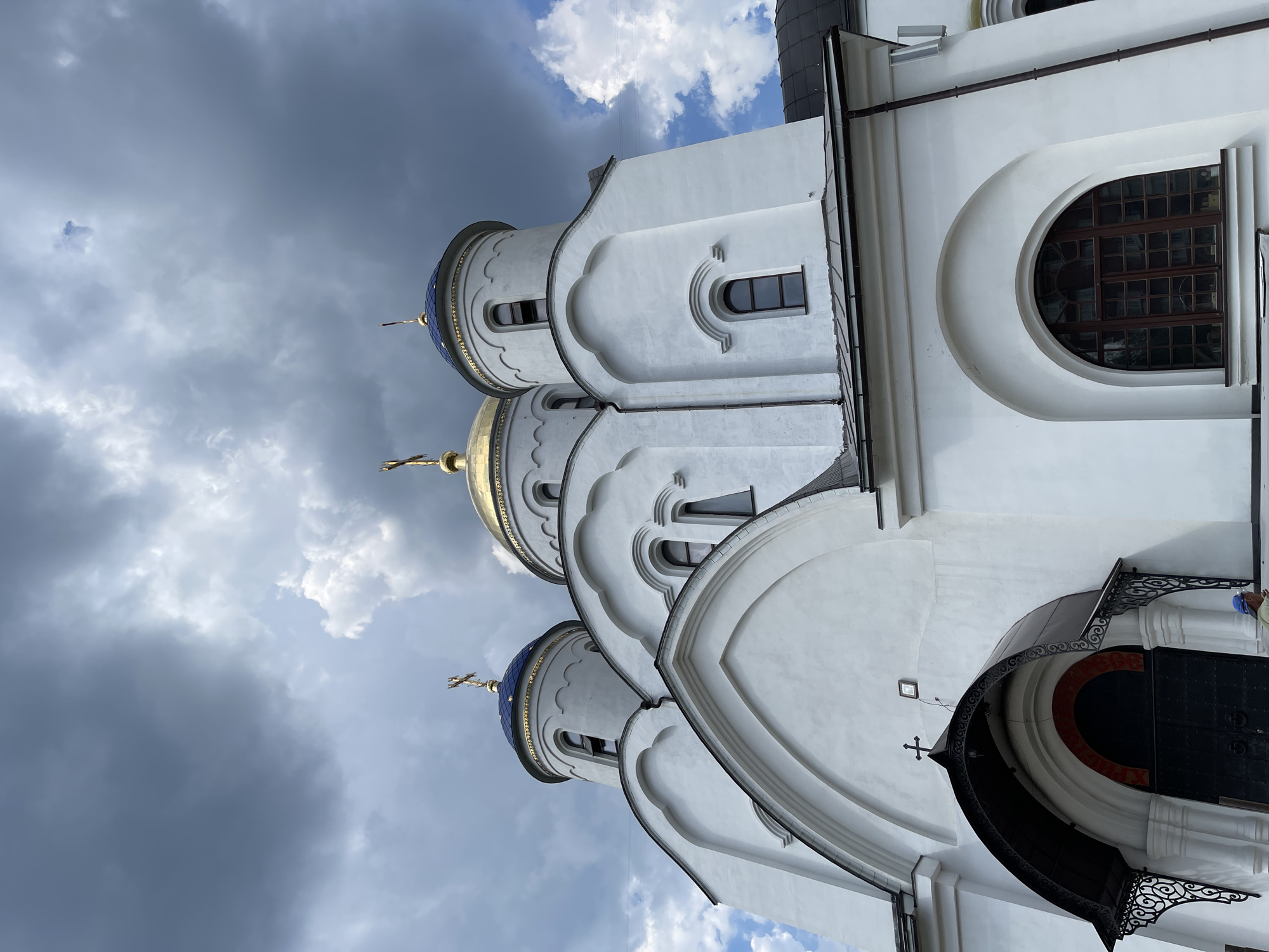 This is a low-angle, eye-level shot of the Church of the Nativity of the Virgin Mary in Staraya Russa, Russia, under a dramatic, cloudy sky. The white facade of the church is prominent, with its distinctive blue domes topped with golden crosses, and a large gold dome in the center. Arched windows and entranceways are visible, as is a dark, ornate canopy over the main entrance. A person in a blue cap is partially visible on the right side of the frame. The clouds in the sky are a mix of dark grey and lighter white, creating a moody atmosphere.