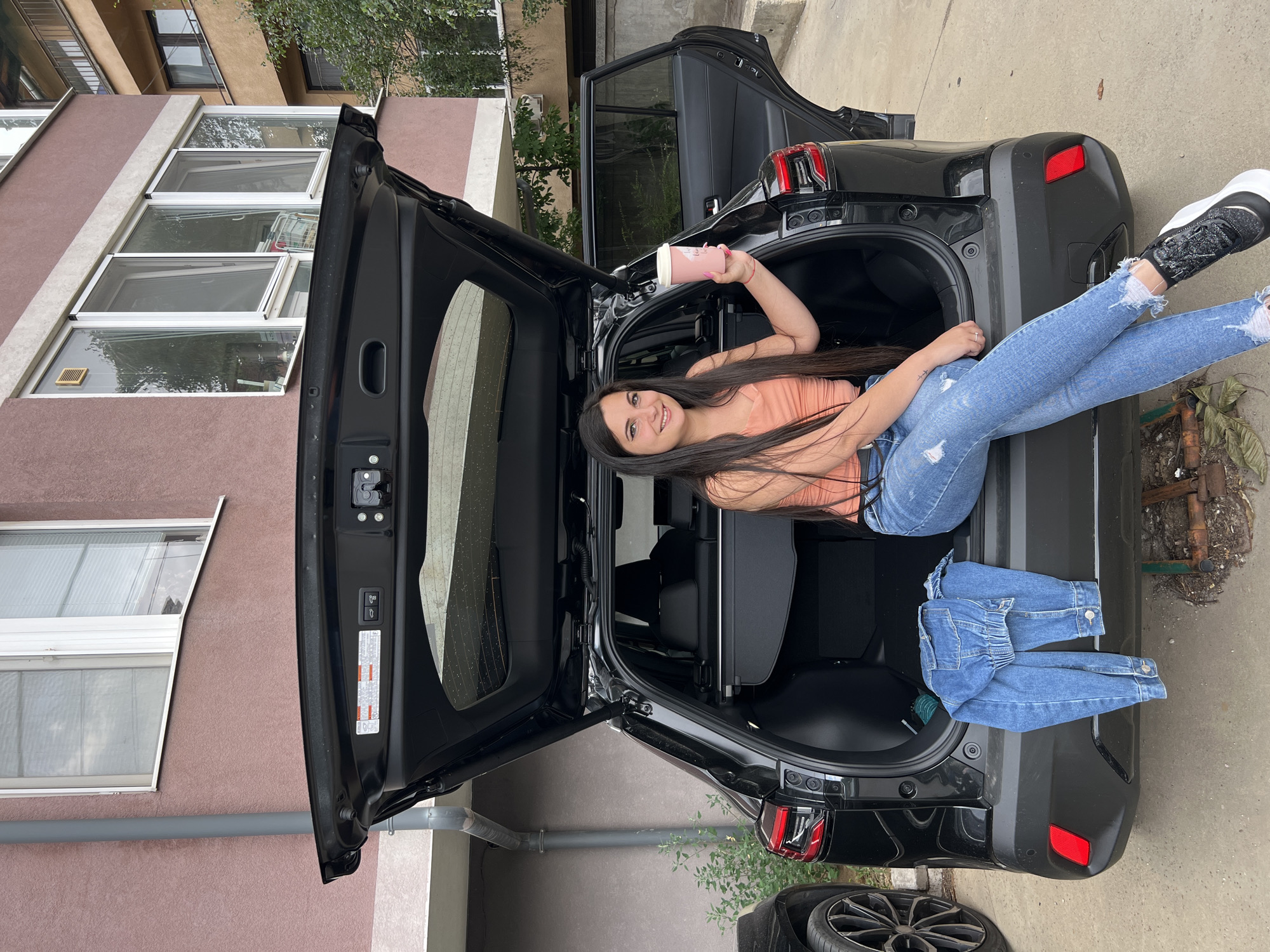 A young woman with long dark hair is sitting in the open trunk of a black car. She is wearing a coral-colored tank top and distressed blue jeans. Her left leg is bent at the knee and resting on the car's interior, while her right leg is extended outward, resting on the car's exterior. In her raised left hand, she holds a light pink cup. A pair of folded blue jeans is placed on the edge of the trunk next to her right leg. The car is parked on a paved surface, with a building visible in the background. The building has a textured, rose-colored facade with several windows. Green foliage can be seen around the building and near the car. The scene appears to be outdoors during the daytime, with overcast weather suggested by the soft lighting. The woman has a relaxed and happy expression on her face. The location is identified as Sofia, Bulgaria.